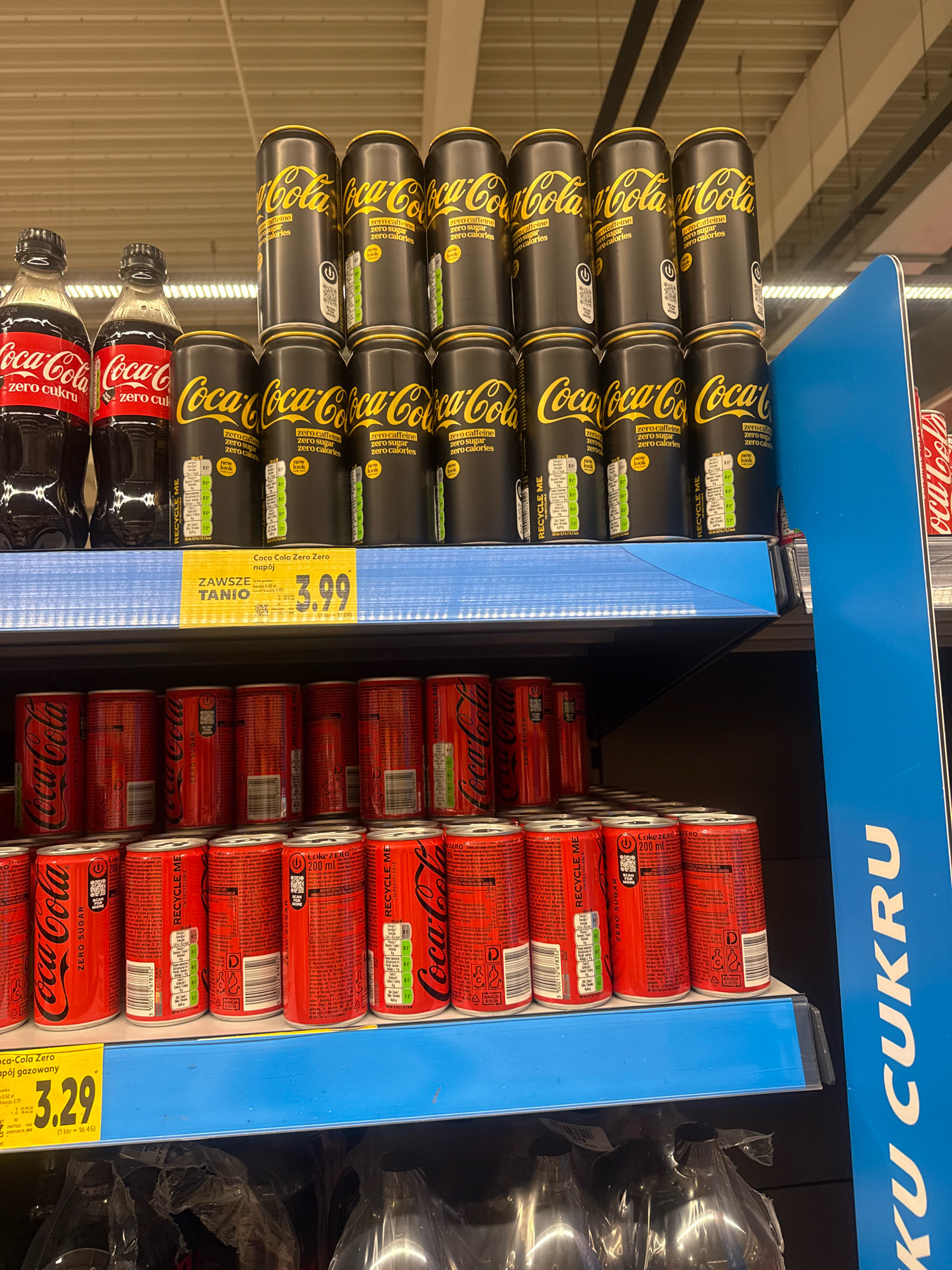 The image displays a retail environment, likely a grocery store aisle, stocked with beverages. The primary focus is on Coca-Cola products, with multiple shelves showcasing cans and bottles. The top shelf features Coca-Cola Zero cans, distinguished by their black and gold packaging, stacked in multiple rows. To the left of these cans are two plastic bottles of Coca-Cola Zero, also in the signature red branding. The middle shelf has more Coca-Cola Zero cans in their red packaging. Below this, the bottom shelf also displays red Coca-Cola Zero cans. A price tag is visible on the middle shelf, indicating "3.99" for "Coca-Cola Zero napój gazowany". A large blue sign with white text "KU CUKRU" is partially visible on the right side of the image. The overall lighting suggests an indoor, well-lit commercial space. The presence of specific product packaging and pricing suggests this is a point-of-sale display. There are no people visible in the image. The location is identified as Gorzow, Poland based on external information.