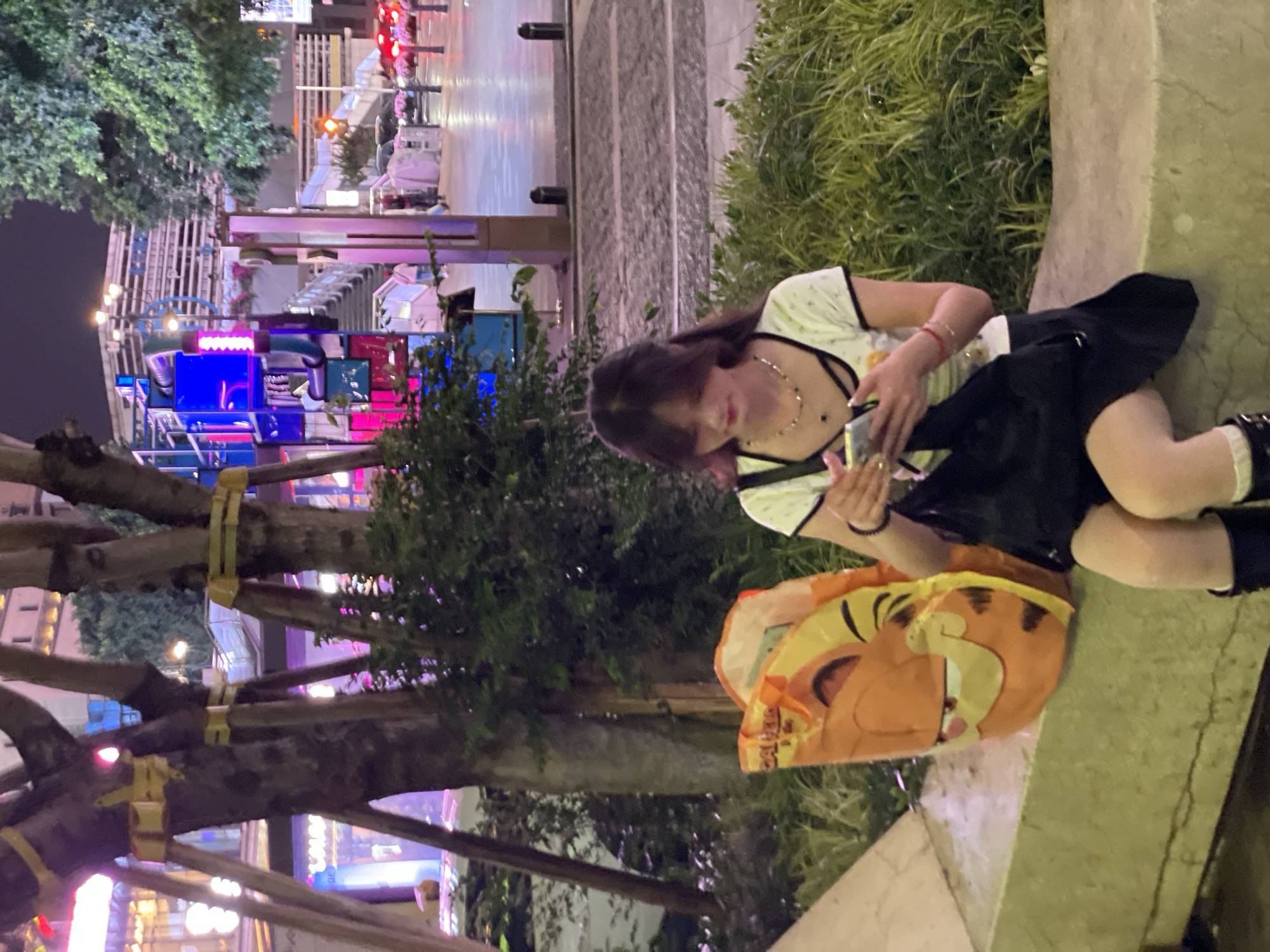 The image captures a young woman seated on a ledge outdoors at night. She has long dark hair and is wearing a light-colored top with dark trim, a dark skirt, and socks with dark shoes. She appears to be looking at a mobile phone she holds in her hands. Next to her is a large, orange-toned shopping bag with a cartoon face. Behind her, there are bushes and trees. In the background are city lights, including what appears to be a colorful light display. The location is Yuzhong District, China.
