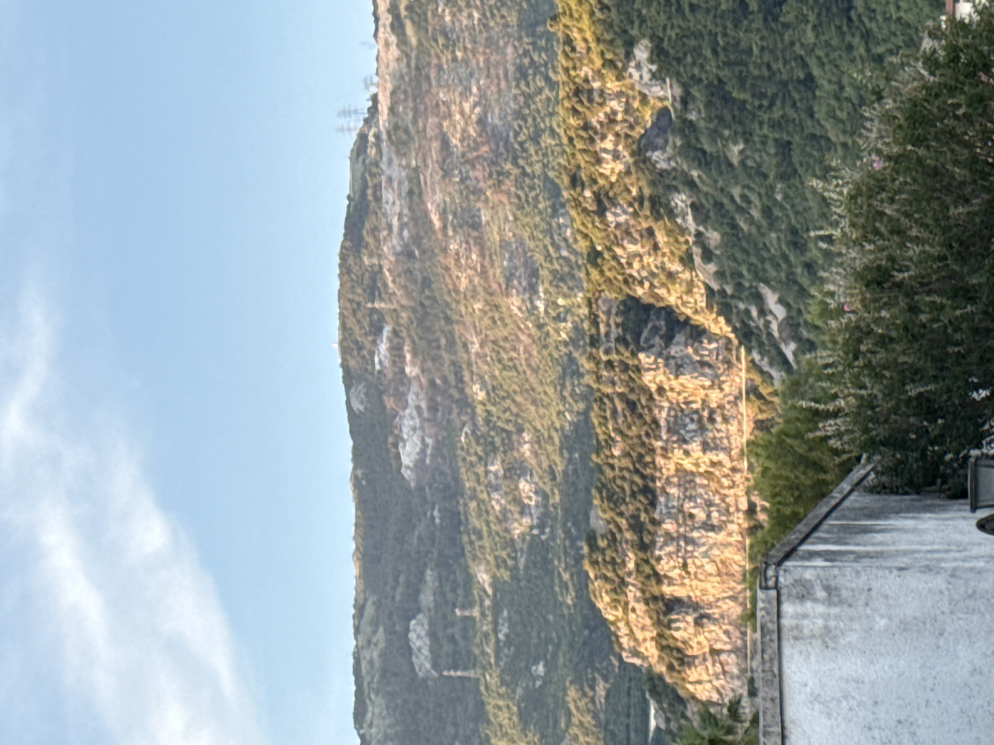 The image shows a mountainous landscape under a clear blue sky with scattered clouds. In the foreground, the corner of a white-washed building is visible, with a dark roofline. Behind the building, lush green trees and vegetation create a dense barrier.
The mid-ground features a dramatic, rugged mountain face illuminated by warm, golden sunlight, highlighting its rocky texture and deep crevices. The mountain is covered in patches of green foliage, suggesting hardy vegetation clinging to the slopes.
Further in the background, a larger, more distant mountain range rises, its slopes covered in a darker green, denser forest. Several tall, skeletal structures, likely electrical pylons, are visible on the crest of this distant mountain. In the middle distance, on the slopes of the mountains, two slender, tower-like structures, possibly old watchtowers or historical markers, are discernible.
The overall scene depicts a natural environment with a touch of human infrastructure, suggesting a location on the outskirts of a populated area, possibly near Budva, Montenegro, given the terrain and the presence of historical or functional structures on the mountains. The lighting suggests it is either early morning or late afternoon.