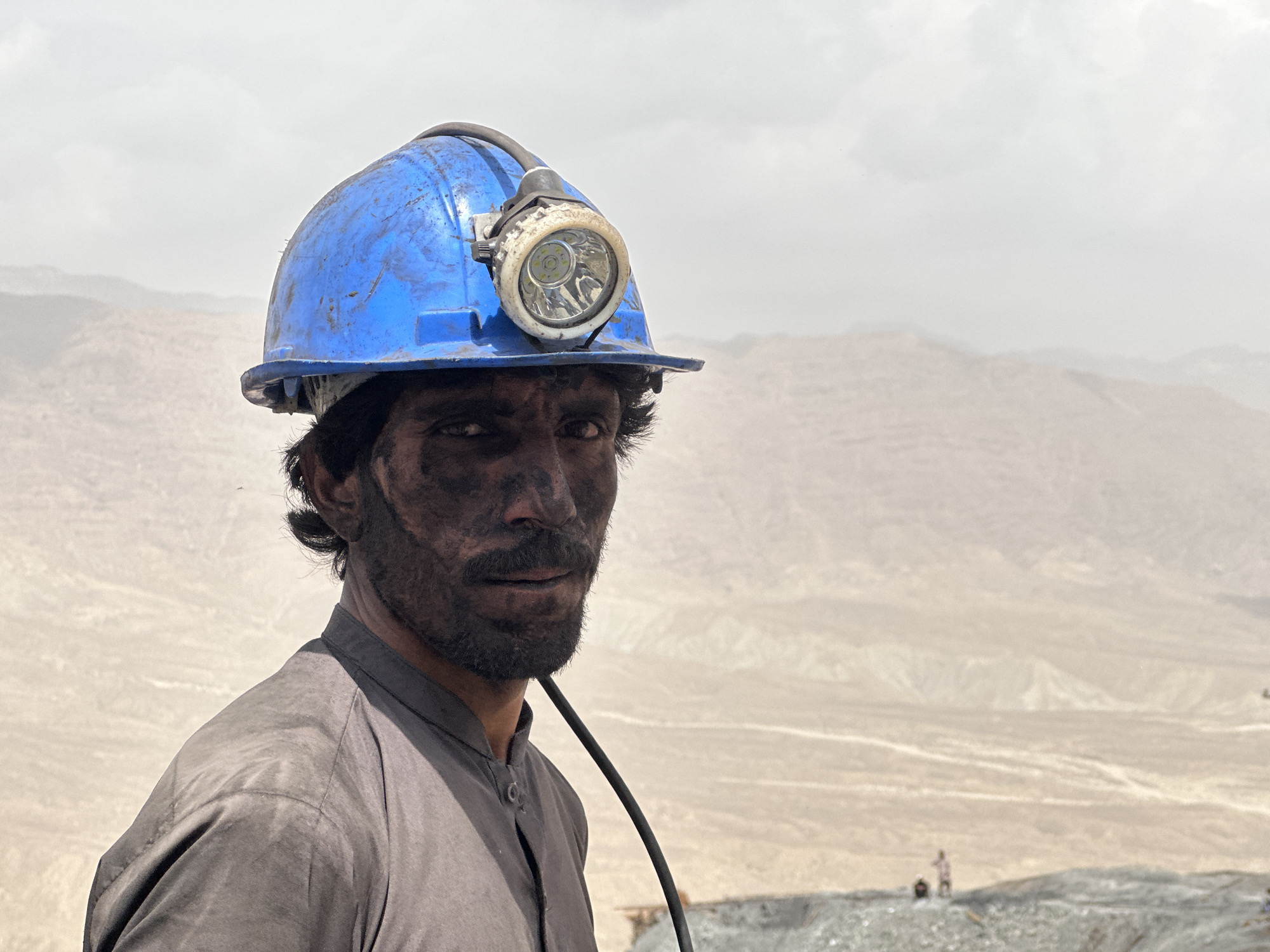 A close-up, medium shot captures a man wearing a dirty blue hard hat with a headlamp. His face is smudged with dark dirt, and he has dark hair and a beard. He is looking towards the right of the frame. He is wearing a dark grey shirt. The background is a hazy, arid landscape with muted brown hills. The man is likely a coal miner in Quetta, Pakistan, a region known for its coal mining industry. The overall scene suggests a difficult and dusty working environment.