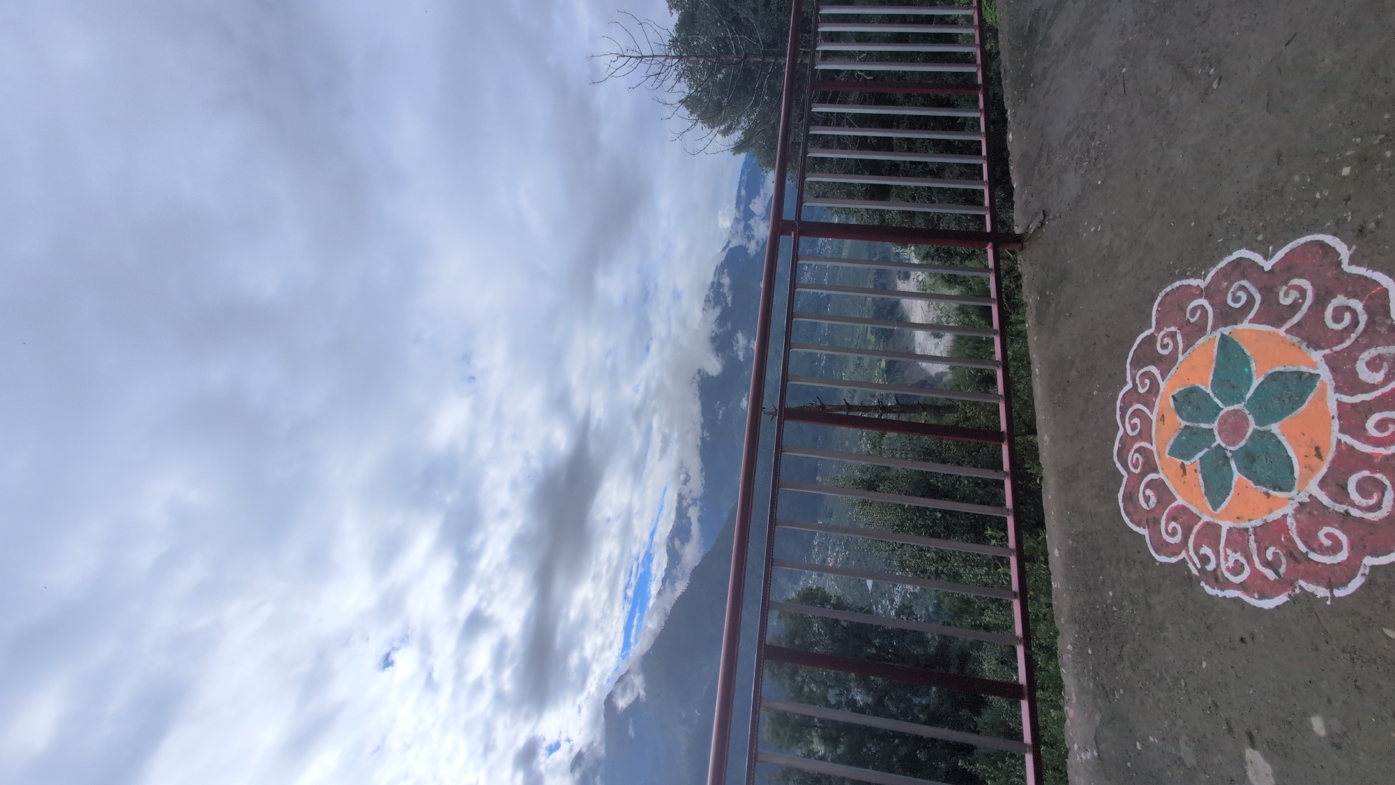 The image depicts a scenic view of a mountainous landscape, likely in or near Pangan, India, under a cloudy sky. In the foreground, a concrete surface features a colorful rangoli, a traditional Indian folk art. The rangoli is circular with intricate patterns in orange, white, and green. Beyond the rangoli, a metal railing with red and white bars separates the foreground from a valley below. This valley contains scattered buildings, indicating a settlement, and a river or water body that appears murky or muddy. Lush green trees and foliage are visible on the slopes of the mountains, which are partially obscured by clouds and mist. The overall impression is one of natural beauty and a glimpse into rural Indian life. There are no people or animals visible in the frame.