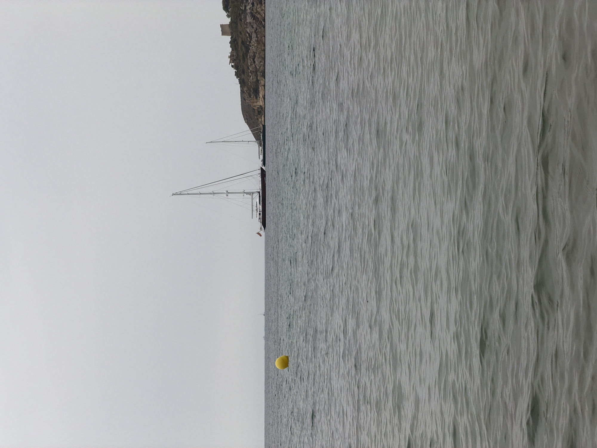 The image depicts a serene seascape with two sailboats anchored in the calm, grey waters. In the foreground, a bright yellow buoy floats, marking the edge of the sea. In the background, a rocky, tree-covered island rises from the water, crowned with a rustic stone tower, likely a historical lookout or fortification. The sky is overcast, contributing to the muted tones of the scene. The overall impression is one of peaceful solitude and natural beauty. The location is the coast of Calvià, Spain.