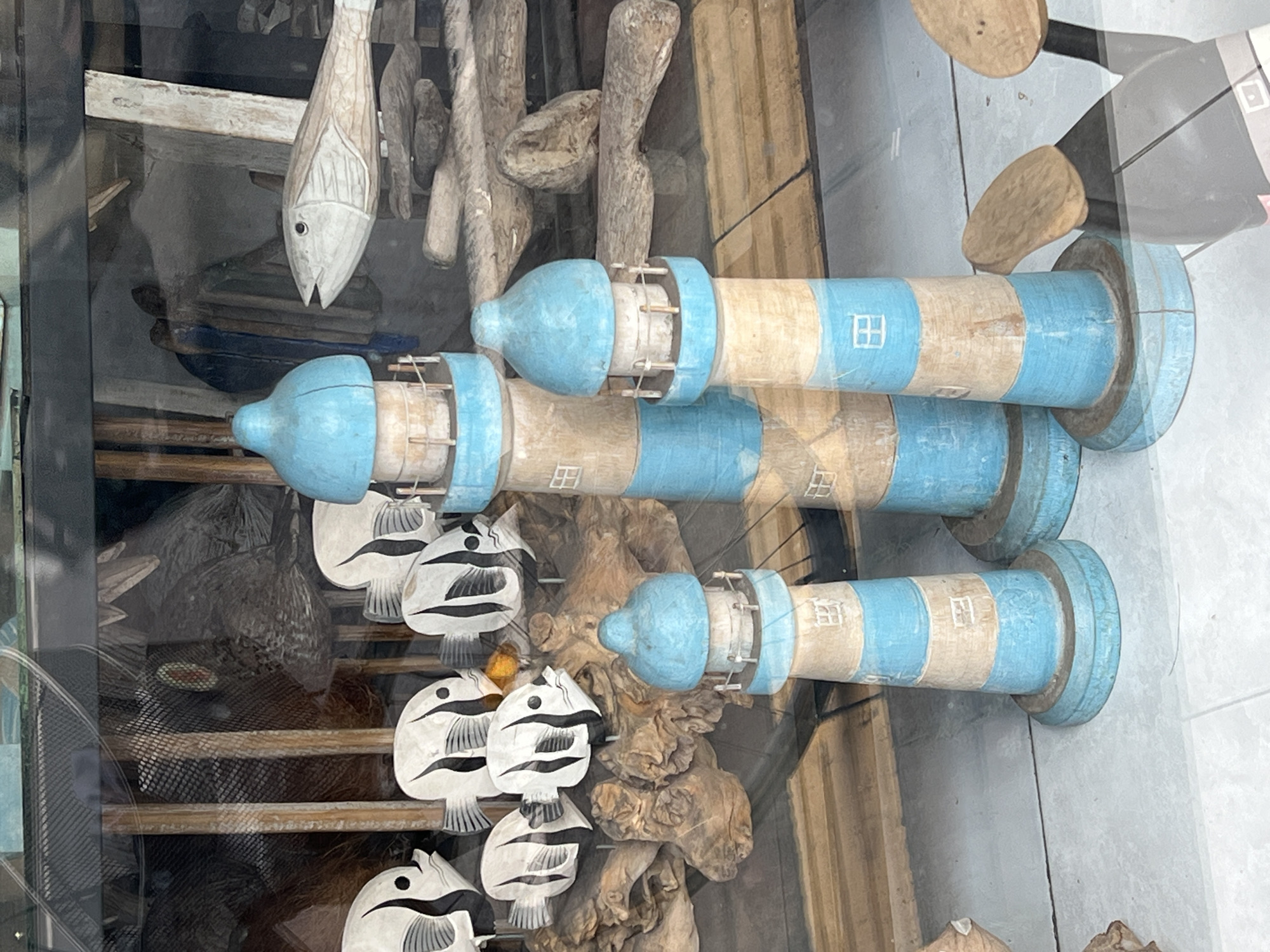 The image depicts a display of nautical-themed crafts in Ubud District, Indonesia. There are three painted wooden lighthouse models, striped in light blue and beige, lying horizontally. The lighthouses have small window-like markings. In the background are wooden fish carvings and other decorative wooden pieces. The display is behind glass, with a slight reflection visible on the surface.