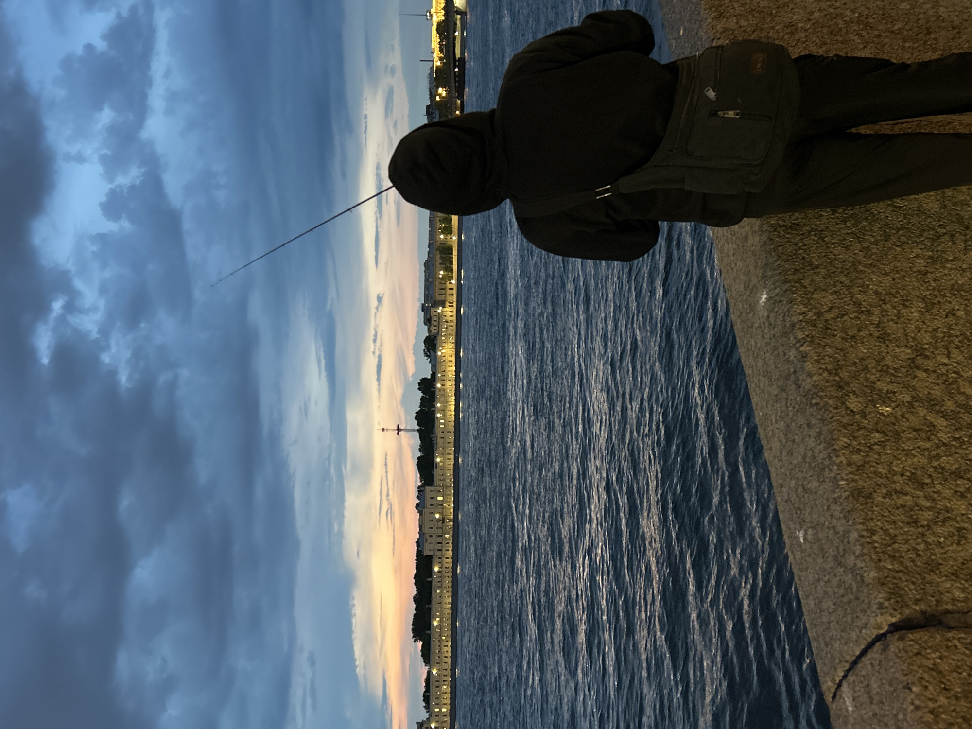The image depicts a solitary individual fishing from a rough-hewn stone embankment at dusk or dawn in the city of Unknown, Unknown. The person is viewed from behind, wearing a dark hooded sweatshirt with the hood up, dark pants, and a dark messenger bag slung over their left shoulder. A fishing rod extends upwards and to the left from their hands. The embankment is illuminated from below, revealing its granular texture and a curved seam on the left. Beyond the embankment lies a wide body of dark blue water, featuring subtle ripples and reflections. Across the water, a distant cityscape is visible, characterized by a long row of illuminated light-colored buildings with numerous glowing windows, backed by dark trees. A tall, slender structure with a crossbar rises above the buildings on the left side of the cityscape. The sky above is heavily overcast with dark blue-grey clouds, exhibiting a band of orange, pink, and yellow light low on the horizon, indicative of sunset or sunrise.