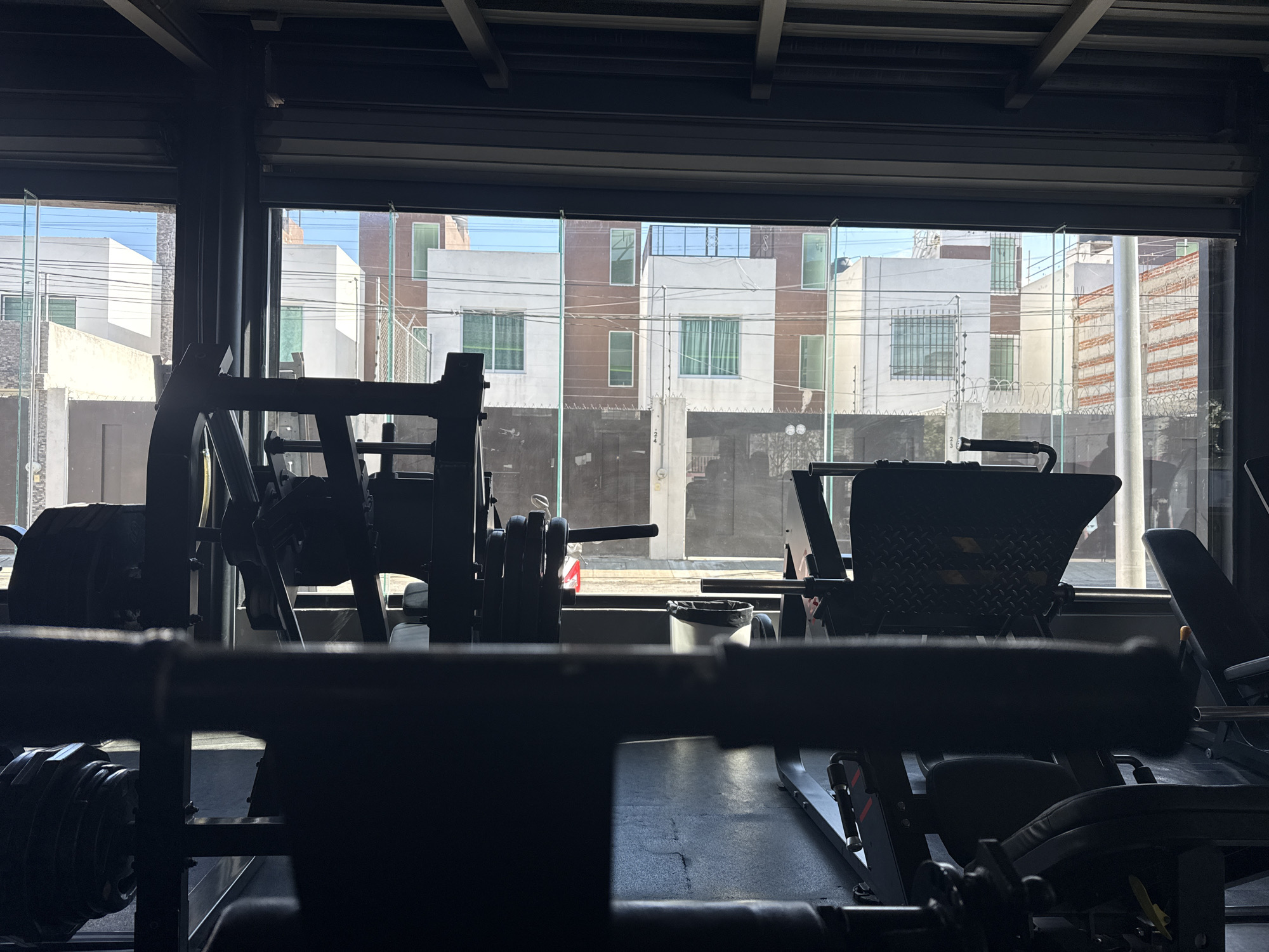 The image is taken from inside a gym located in Puebla, Mexico, with a large window offering a view of the street and buildings opposite. Gym equipment is silhouetted in the foreground, including leg press machines and weightlifting apparatus. The focus is drawn to the exterior scene through the window, which depicts residential buildings in varying states of repair, some with white facades and others with brown accents. Security features like fencing and barbed wire are visible on some properties, suggesting an urban environment. The sky is clear and blue, indicating a sunny day. A trash can is visible next to the leg press machine.