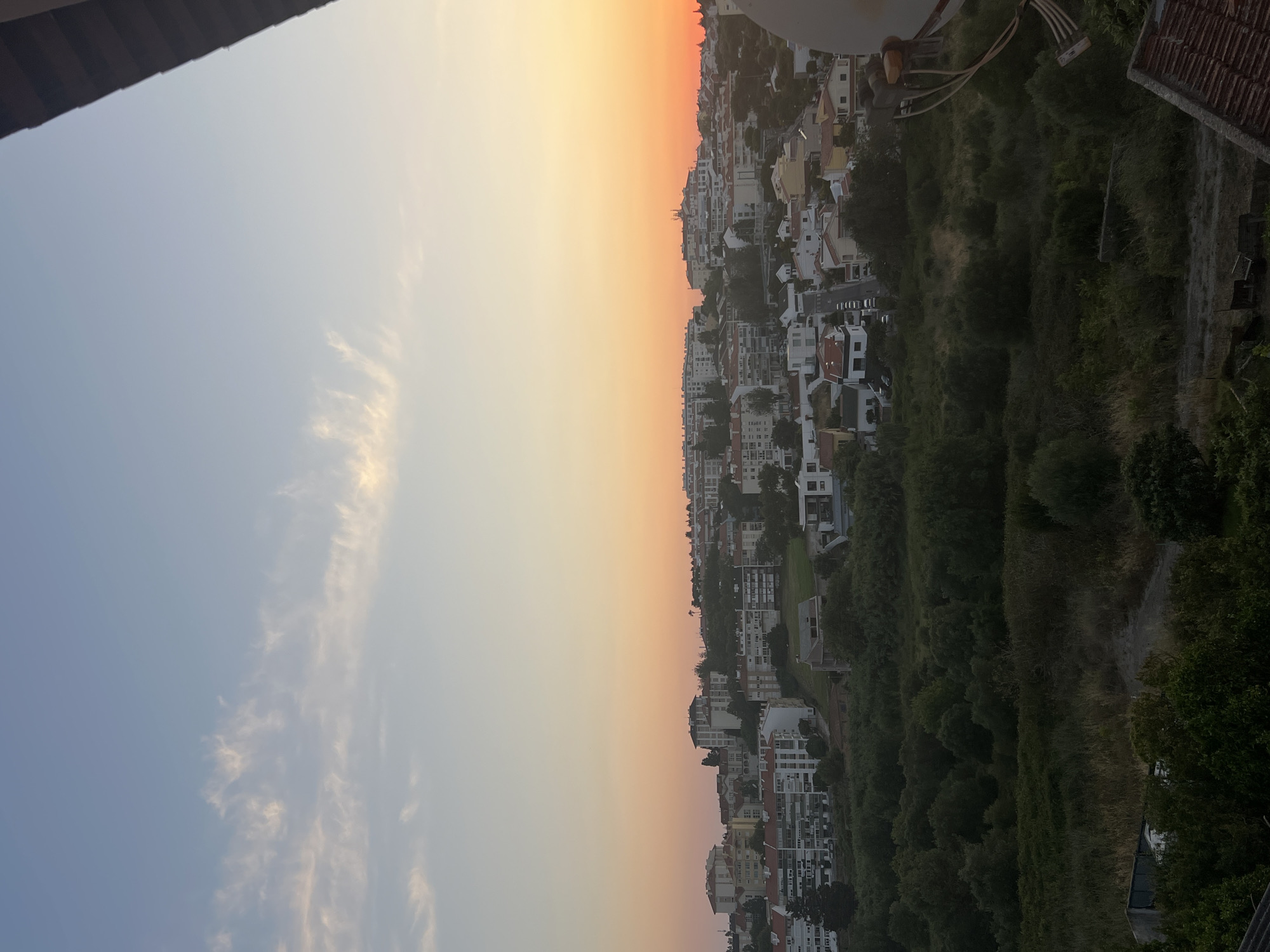 An exterior, eye-level, full shot captures a panoramic view of the city of Cascais, Portugal, during a vibrant sunset. The sky transitions from a pale blue at the top to a gradient of soft orange and yellow hues near the horizon. Wispy white clouds are scattered across the upper portion of the sky.
The city itself is built on a hillside, with numerous multi-story buildings featuring white facades and red-tiled roofs. These structures are densely packed, creating a cityscape that stretches across the middle ground. Vegetation, consisting of lush green trees and shrubbery, covers the lower half of the image, acting as a natural foreground that separates the viewpoint from the city.
A residential street, lined with parked cars, winds its way through the urban landscape. In the lower right quadrant, a portion of a satellite dish is visible, suggesting proximity to residential areas. The overall atmosphere is serene, depicting a typical evening in a coastal Portuguese city.