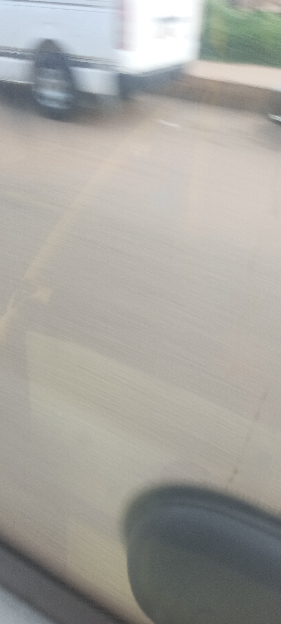 The blurred image depicts an outdoor scene, likely captured from inside a moving vehicle in Bukuru, Nigeria. A white vehicle, possibly a truck or van, is partially visible on the left side, exhibiting significant motion blur, particularly around its wheels. The dominant feature of the photograph is the road or street surface, which occupies most of the frame and shows horizontal streaks indicative of speed and movement. In the upper right corner, indistinct green blurs suggest roadside vegetation. A dark, rounded, blurry object in the bottom right corner strongly resembles a car's side-view mirror, confirming the perspective from within a moving automobile. The scene conveys a sense of transit during daylight hours. No specific time of day, weather conditions, or text are clearly discernible.