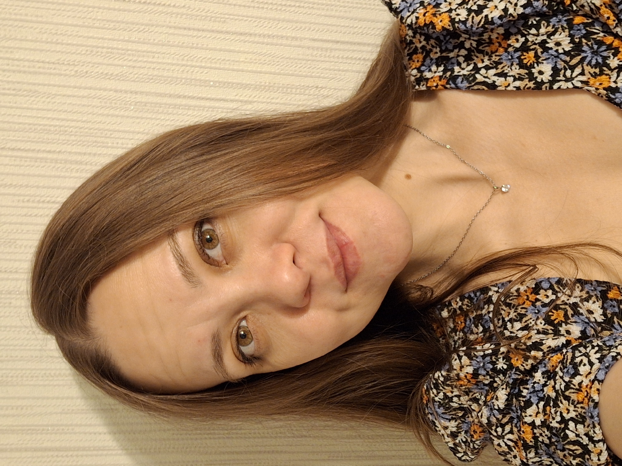 A woman with brown hair is lying down on a textured cream-colored surface, possibly a bed or couch. She is wearing a floral-print top in black, blue, and orange. A silver necklace with a small pendant is visible around her neck. She is looking at the camera with hazel eyes. The background is out of focus.