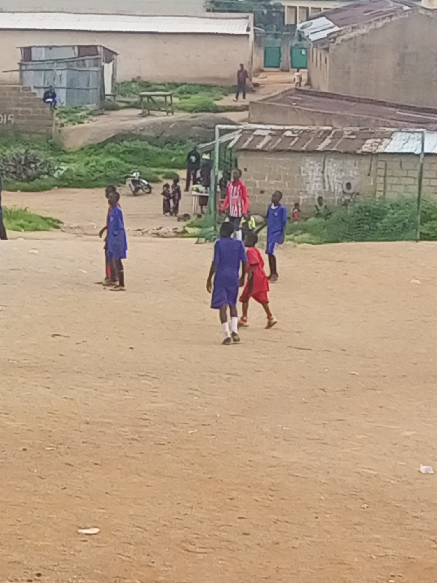 In Jos, Nigeria, a group of children are playing soccer in a dusty, open field. The area is characterized by its unpaved ground and a scattering of modest, tin-roofed structures that appear to be residences or workshops. In the background, more substantial buildings with corrugated metal roofs are visible, along with some greenery and a hint of a fence. The children are dressed in athletic wear, some in blue and others in red and white striped shirts. One boy in a blue jersey is captured mid-stride, his attention focused on the game. Another, in red shorts and a red shirt, is looking away from the viewer. A third boy in a blue outfit stands still, while a fourth, also in blue, is partially obscured from view. The overall scene depicts a typical informal gathering and recreational activity in a developing urban environment.