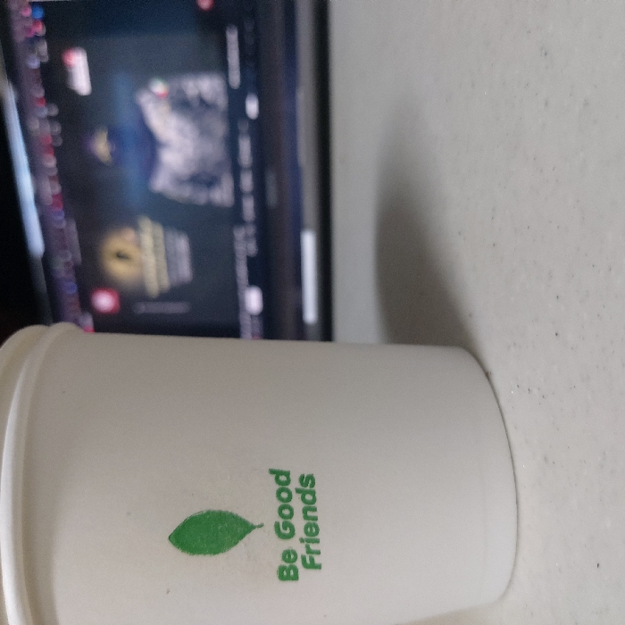 A white disposable paper cup, adorned with a green leaf logo and the phrase "Be Good Friends," rests on a light-colored, speckled surface in 김포시, South Korea. In the blurred background, a laptop screen displays a figure wearing a dark hood and a camouflage-patterned jacket, alongside a golden emblem and obscured text. The scene implies an indoor setting, likely a desk or table, with the cup positioned in the foreground and the laptop screen providing ambient light. No individuals are visible.