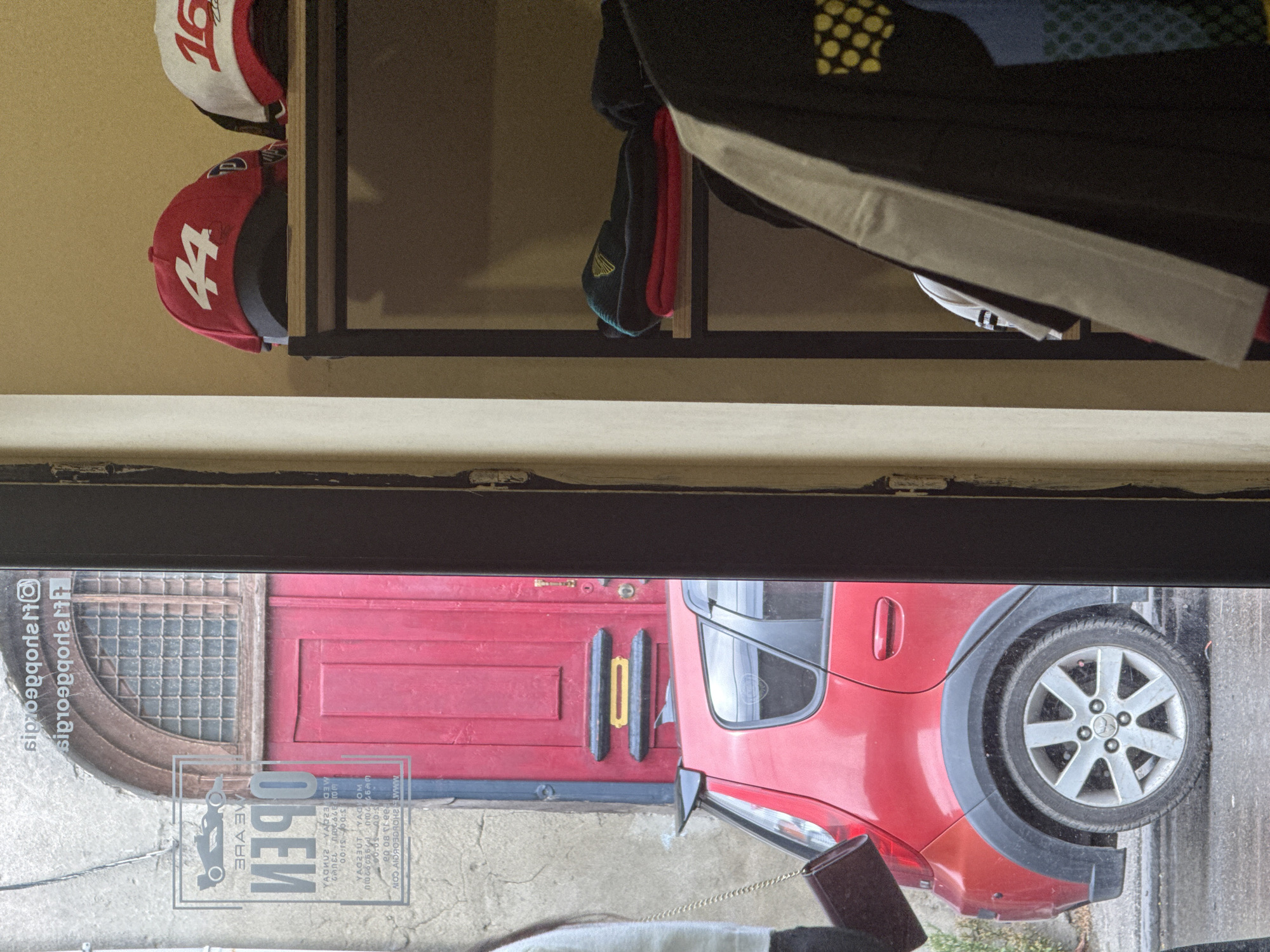The image shows a view through a glass door into a shop, with a red car parked outside. The shop appears to be a retail store selling clothing and accessories, with shelves of hats and jackets visible. A red door and an arched window with a grid pattern are visible through the glass. A woman is partially visible on the left side of the image, holding a purse. The overall scene suggests a street-level retail establishment.