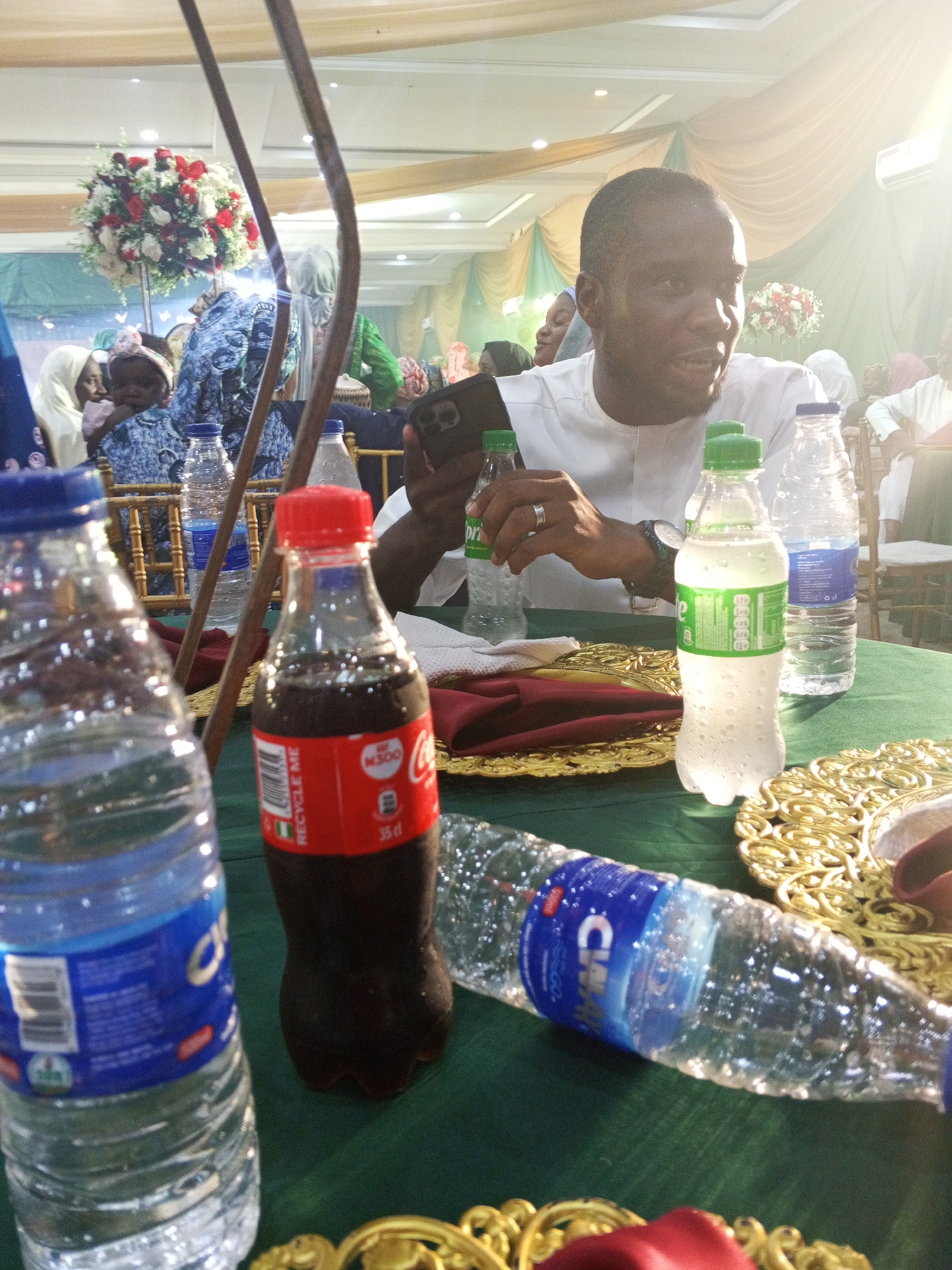 A man in a white tunic is seated at a table, holding a smartphone and looking to his right. In front of him are several bottles of beverages, including Coca-Cola and bottled water. The table is covered with a green tablecloth, and ornate gold platters are visible. In the background, the scene appears to be an indoor event, possibly a wedding reception or a banquet, with draped curtains, floral arrangements, and other guests. The lighting is bright, with a spotlight effect coming from the top right of the image. The man is wearing a ring on his left hand and a watch on his left wrist. The overall ambiance suggests a celebratory gathering.