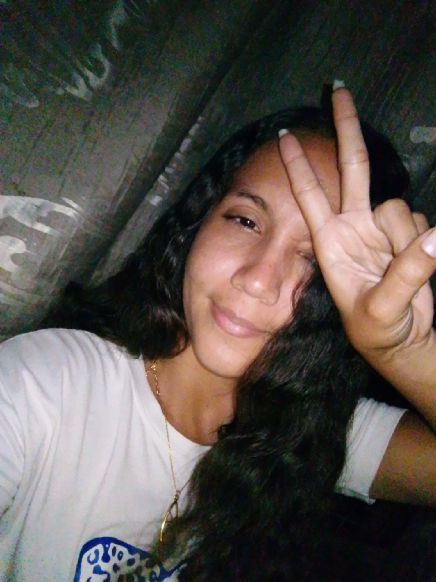 A woman is posing indoors, smiling and giving the peace sign. She has long, dark, wavy hair and is wearing a white t-shirt and a gold necklace. The background appears to be some type of textured wall or fabric. This photograph was taken in Juan Griego, Venezuela.