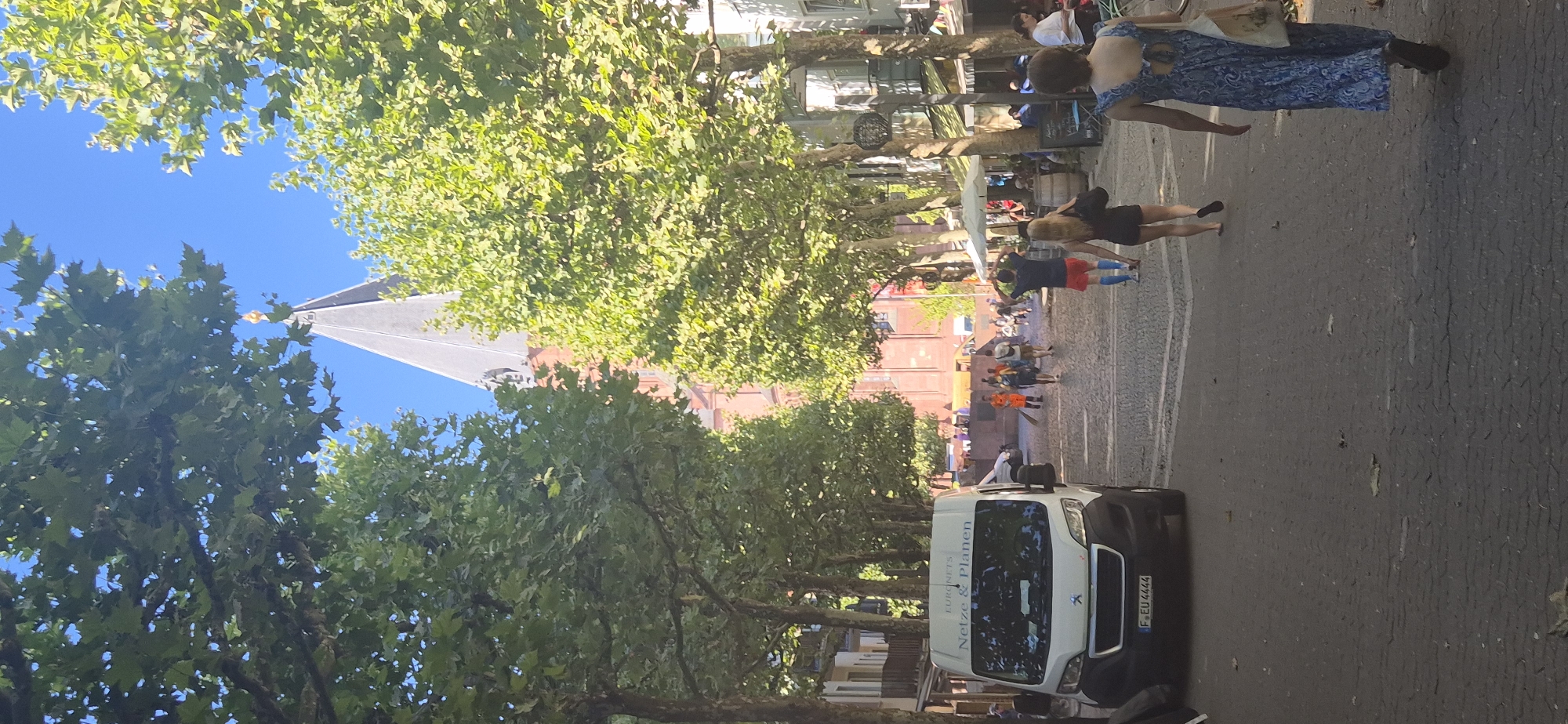 In Mainz, Germany, a sunny street scene unfolds, featuring a white van parked on the left with the text "Euronets Netze & Planen" visible on its side. The van has a license plate reading "F EU 4444". The street is paved with cobblestones and lined with mature trees that cast dappled shadows. In the background, a tall brick building with a pointed, dark-tiled spire topped with a golden finial dominates the view. Several people are walking on the street, some in casual attire, including a person in an orange uniform sweeping the area. Further down the street, outdoor seating areas for cafes are visible, with umbrellas providing shade. The sky is a clear blue.