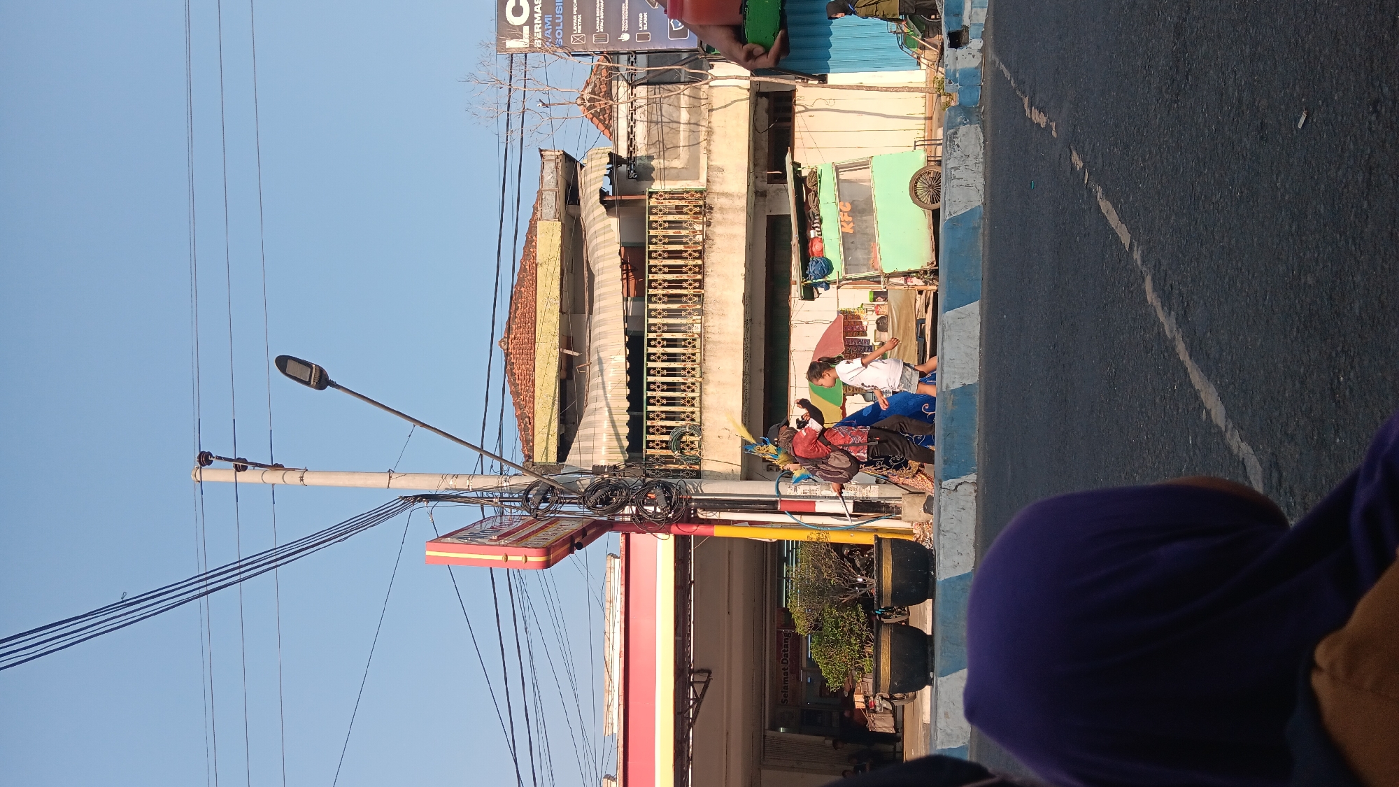 The image captures a street scene in Tanjung Anom, Indonesia, under a clear blue sky. In the foreground, a person wearing a purple headscarf is partially visible, facing away from the camera. The midground shows a street with a blue and white striped curb. On the sidewalk, a woman in a red and black patterned shirt and dark pants appears to be interacting with a young girl in a white t-shirt and shorts, who is walking away from the camera. Behind them is a light green food cart with the "KFC" logo. To the left of this scene, a red and yellow sign with text is visible, attached to a building with awnings and a balcony. Electrical wires and a streetlamp are prominent in the sky. A building with a tiled roof and a second floor balcony with an ornate railing stands behind the food cart and the people. The environment appears to be a commercial area with a gas station visible to the far left.