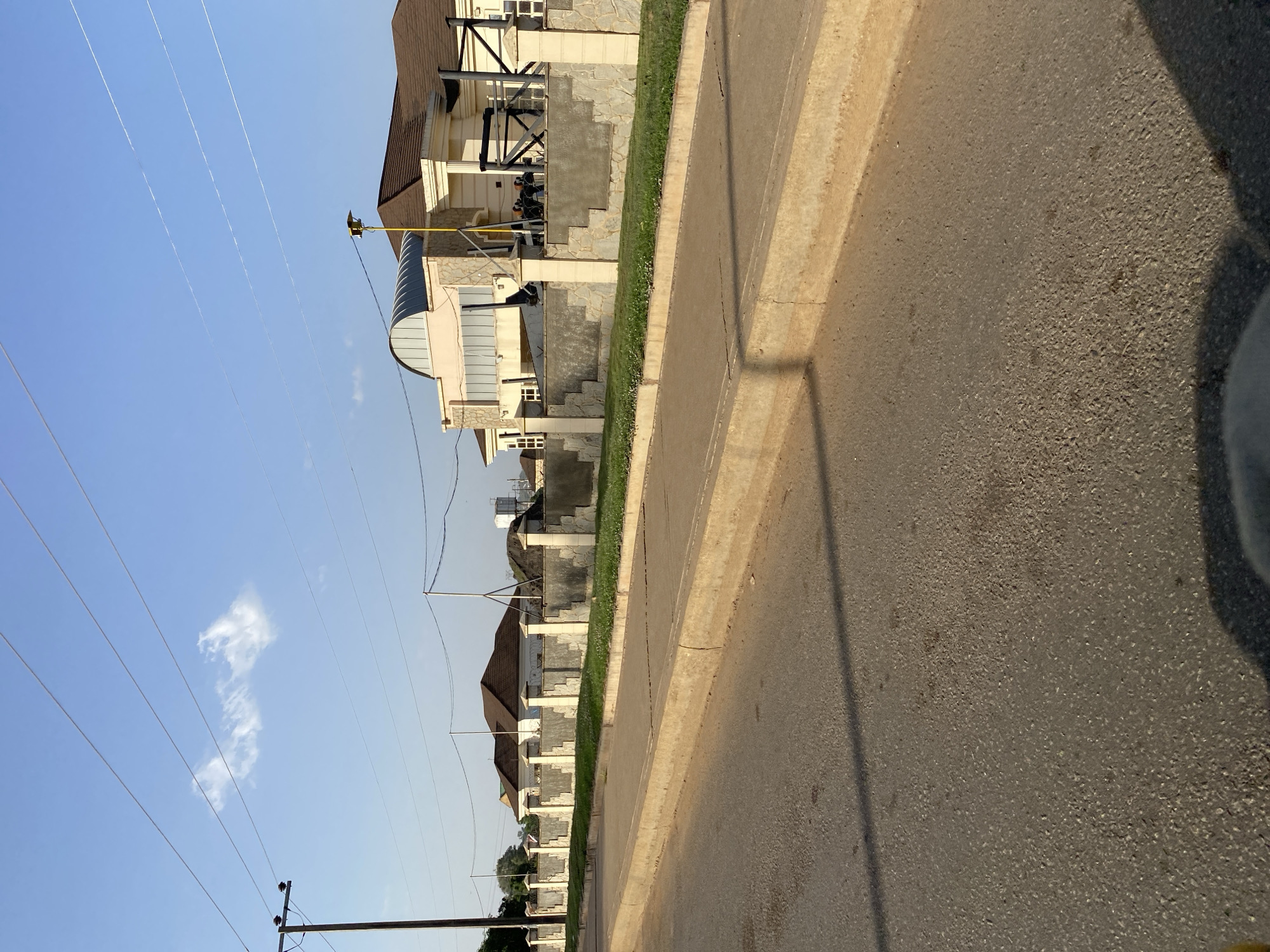 The scene depicts a street in Bukuru, Nigeria. The view is from street level, looking along a row of residential buildings lining the left side of the street. A paved road runs along the right side of the image. There's a curb separating the road from the landscaped area along the buildings. Utility poles and power lines are visible against a clear, blue sky with some clouds. No people are visible in the scene.