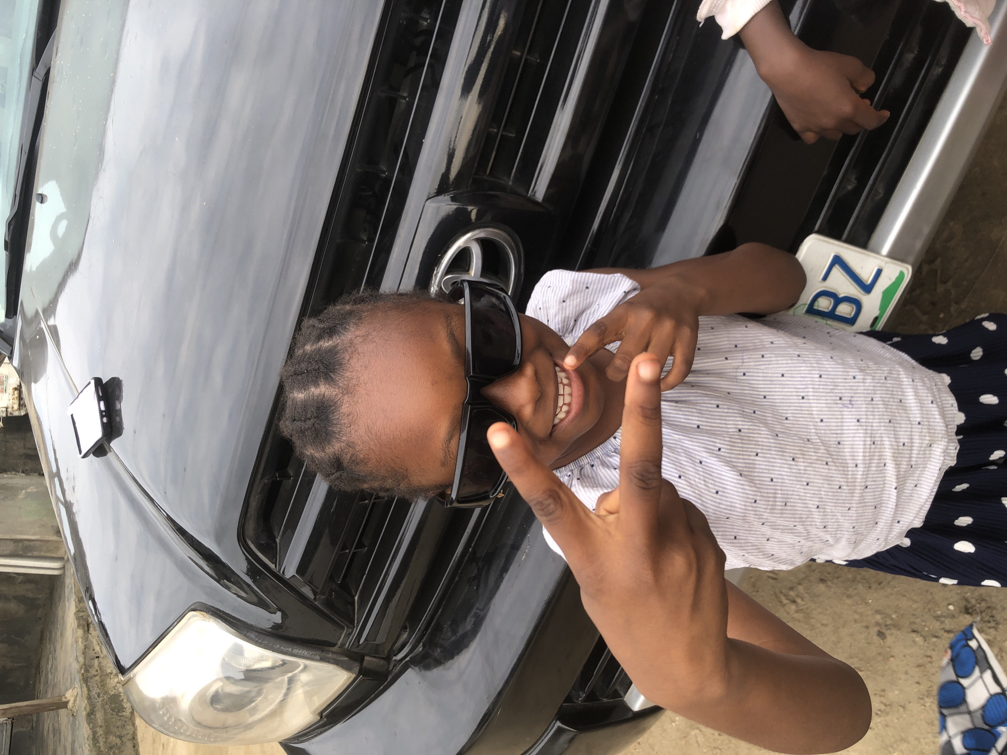 The image captures a close-up, eye-level shot of a young Black girl standing in front of a black SUV. She is wearing large, dark sunglasses and a white and blue striped shirt with a polka dot skirt. She is making a hand gesture, with her left hand held up, fingers spread to form a peace sign, and her right hand's index finger pointing towards the camera. The front of the black SUV, including its grill and headlight, is visible behind her. A white license plate with the letters "BZ" in blue is attached to the front bumper. The ground is covered in sand and gravel. The scene appears to be outdoors, likely in Ado, Nigeria, given the context provided.
