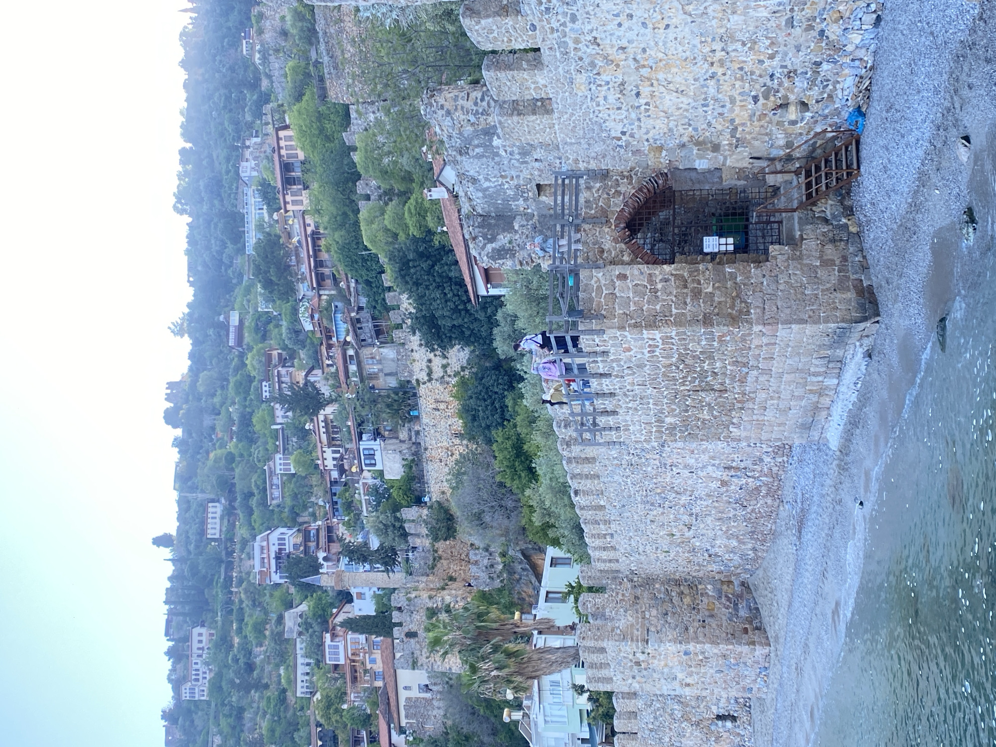 The image captures a scenic view along the historic stone walls of Alanya, Turkey, overlooking the coastline and a verdant, built-up hillside. The perspective is from alongside the robust stone fortifications, which run parallel to a pebble beach and the calm, clear sea.
Several people are visible on the stone wall, appearing to observe the landscape. One individual wears dark clothing with a pink bag on their arm, while another is in a light top. A rustic metal ladder extends from the wall down to the beach, and nearby, a grated metal structure or gateway is integrated into the wall at the water's edge. A blue plastic bag is noticeable on the pebbly shore near the ladder. Behind the metal grate, a green bin and an unreadable white sign are faintly visible.
The steep hillside behind the walls is densely covered with trees and numerous buildings, ranging from residential houses to larger structures, ascending towards a distant summit where another fortification or castle element is present. A minaret is discernible among the buildings on the slope. The lighting is soft and somewhat hazy, indicating either early morning or late afternoon, with a pale sky.
