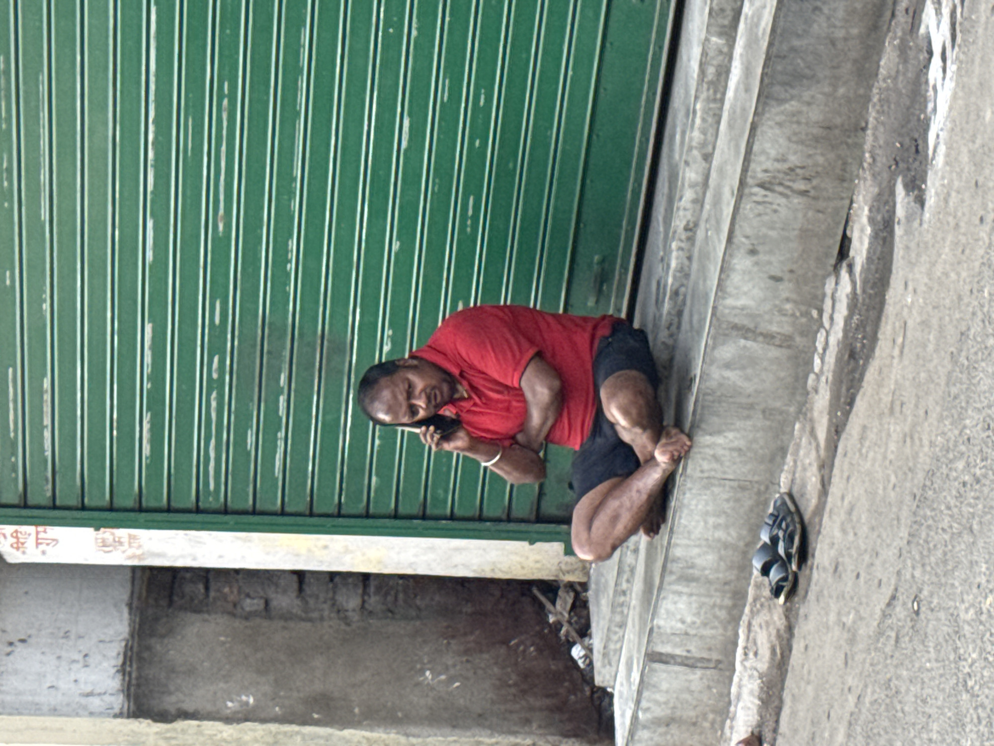 A man wearing a red shirt and dark shorts sits barefoot on a concrete ledge next to a closed green corrugated metal door. He is holding a cell phone to his ear, seemingly engaged in a call. His discarded sandals sit on the pavement next to him. The scene appears to be outdoors, possibly near a storefront in Kolkata, India. The surroundings include a gray concrete surface and a weathered building facade.
