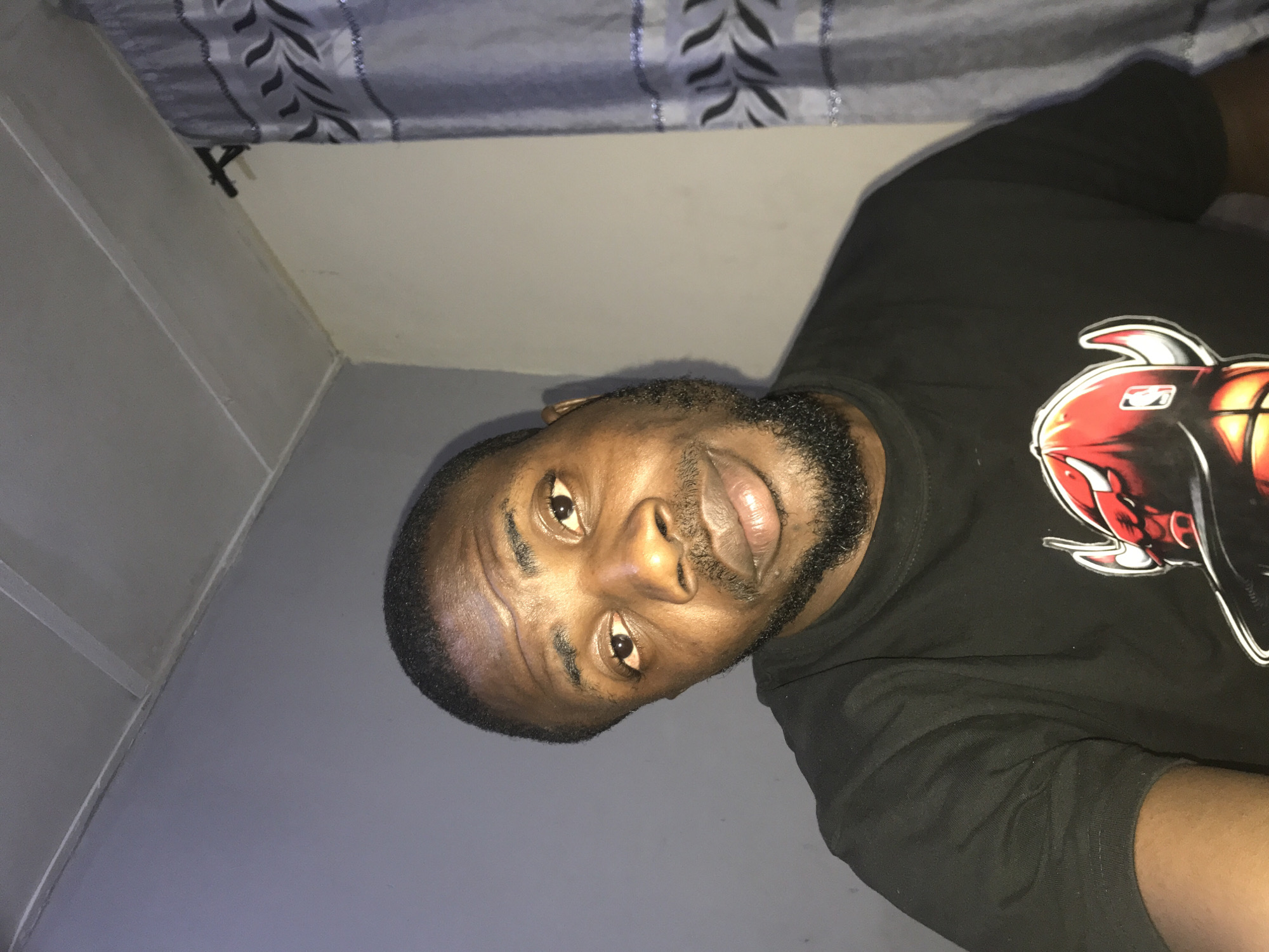 A dark-skinned adult male, approximately 30-40 years old, with short dark hair and a trimmed beard and mustache, looks directly at the camera with a neutral expression. He is wearing a short-sleeved black t-shirt featuring a large graphic of a red Chicago Bulls logo, depicting a bull's head wearing a baseball cap with an NBA emblem, situated above a basketball pattern.
The background shows an indoor setting. To the subject's left is a solid light gray wall. To his right, a light beige wall meets the gray wall at a corner. The ceiling, visible in the upper portion of the frame, is white and constructed of rectangular panels with visible seams. On the right side of the image, a light blue-gray translucent curtain with a repeating black abstract leaf or wave pattern is visible, hanging from a dark curtain rod. Artificial overhead lighting illuminates the scene. The location is in the city of Unknown, Unknown.