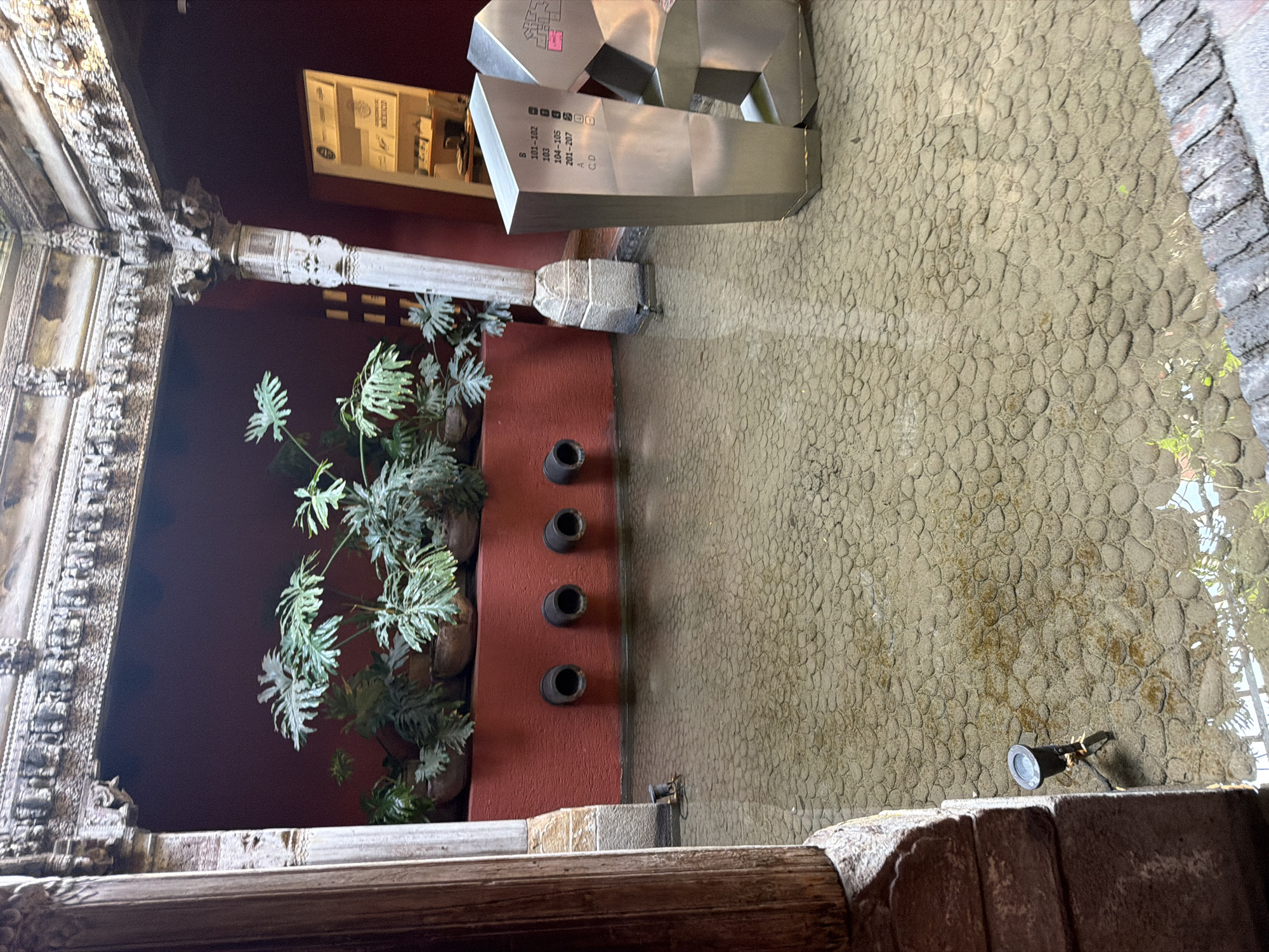 The image depicts a courtyard setting, presumably in Puebla, Mexico, given the presence of colonial architecture elements. The courtyard is characterized by a textured, cobblestone-like ground surface. The view looks up and to the right to display the vertical elements of the courtyard.
On the left, there is a decorative, carved stone architectural detail, possibly part of a window frame or a column. Adjacent to this is a dark red or maroon wall with multiple potted plants arranged vertically. The plants appear to be of a broad-leafed, possibly tropical variety. Additionally, a row of four dark-colored cylindrical objects, maybe drainage spouts or decorations, are embedded within the dark red wall. Above, a large, modern metallic sign displaying room numbers is present.
The scene appears to be a blend of traditional architectural elements and modern signage or decor. The environment suggests a mixture of natural and artificial elements, with plants juxtaposed against stone and modern metal.