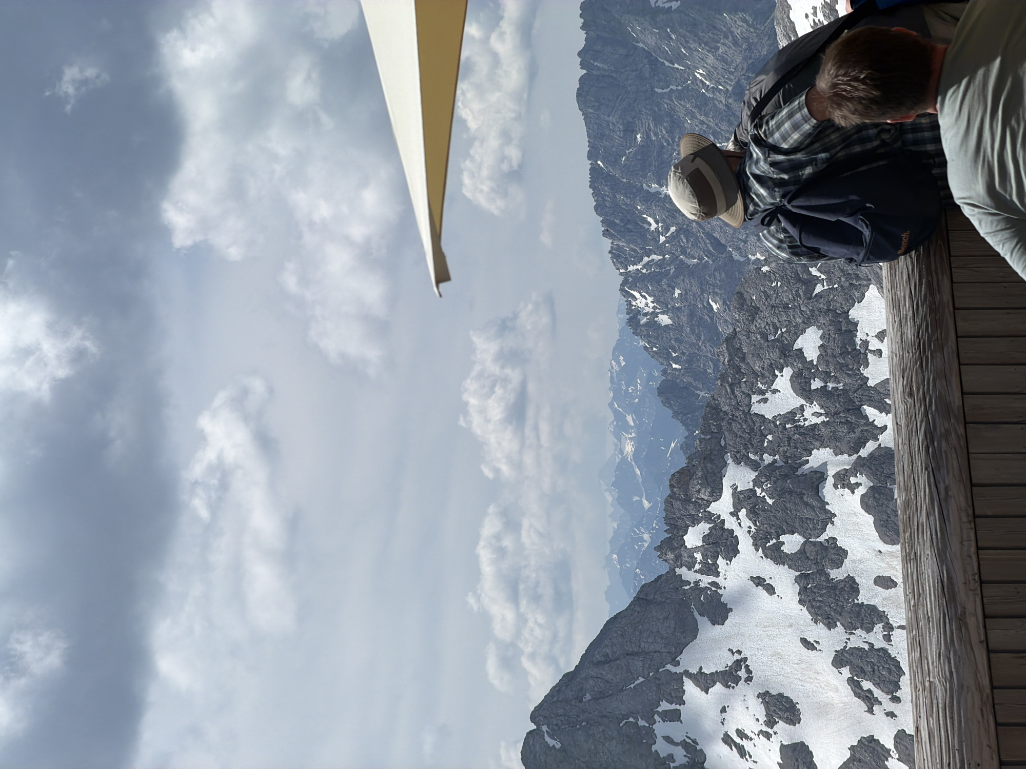 The image shows a view of the Zugspitze mountain range, likely from the viewing platform at the AlpspiX or a similar vantage point near Garmisch-Partenkirchen, Germany. The foreground features a wooden railing and a dark wooden fence. Two individuals are seated, facing away from the camera and towards the mountains. The person on the left wears a hat, a plaid shirt, and a backpack. The person on the right wears a grey t-shirt. The background is dominated by a vast mountain landscape with snow-capped peaks and rocky outcrops, under a sky with scattered clouds. An off-white umbrella is partially visible in the upper right corner.