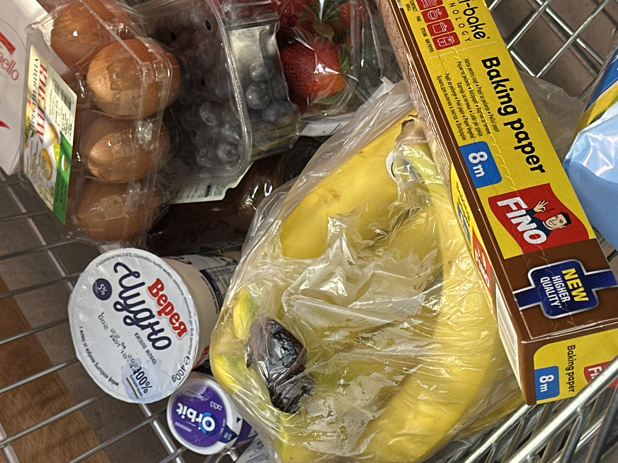 The image shows a shopping cart containing various food items.
The food items include: a package of eggs, a container of blueberries, a container of strawberries, a package of bananas with a dark spot/damaged banana, a container of yogurt ("Верея Чудно") with text in Bulgarian, and a package of "Orbit" gum. There is also a roll of "Fino" baking paper. The cart appears to be located in a grocery store or supermarket, implied by the items purchased. There are no people visible.
Given the presence of Bulgarian text on the yogurt and the gum, this image was likely taken in Bulgaria. Without other location clues within the image, it is not possible to determine if it was specifically taken in Varna.