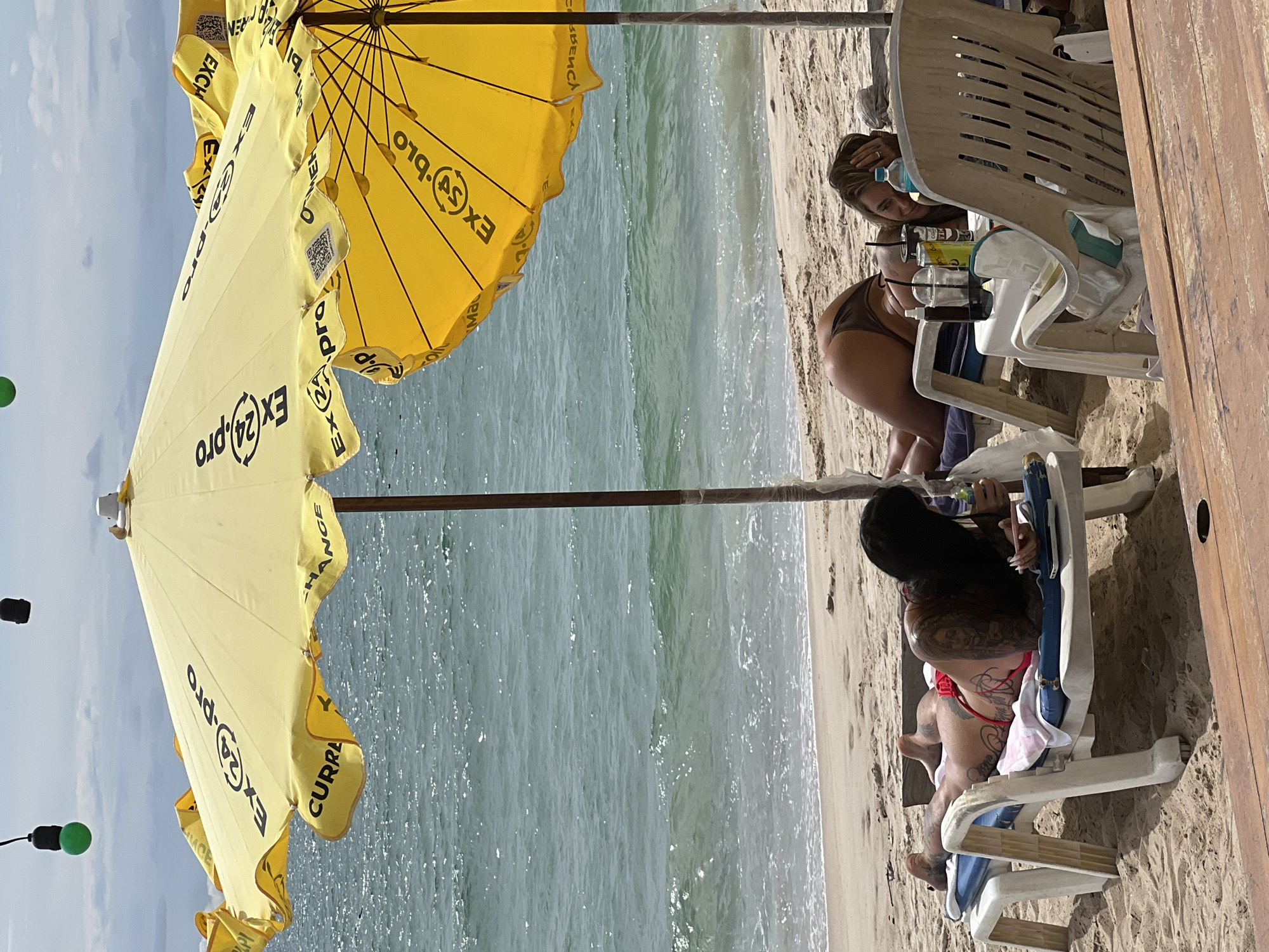 Two women are sunbathing on lounge chairs on a sandy beach. The woman on the left is lying on her stomach, wearing a red bikini bottom and is heavily tattooed. She is holding a phone and a drink. The woman on the right is also lying on her stomach, wearing a brown bikini. She is looking towards the camera. Both women are under a large yellow umbrella that has "EX24.pro" and "CURRENCY EXCHANGE" written on it. The ocean is visible in the background. There are other lounge chairs and a table with drinks in front of the woman on the right. The scene appears to be in a tropical or warm climate.