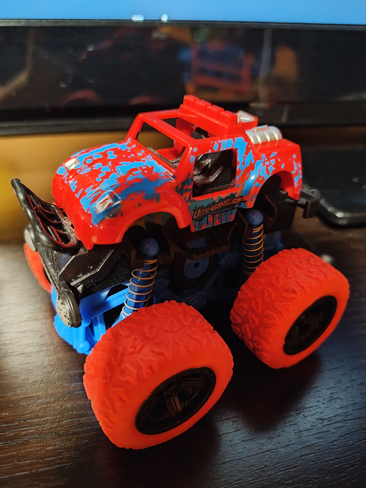 A red and blue toy monster truck sits on a dark, wooden surface. The truck has large, orange wheels with black rims. It is decorated with abstract blue splatters on its red body. The truck's suspension system is visible, featuring yellow springs. In the background, a blurred screen or monitor is present. The image is taken from a low angle, focusing on the toy.