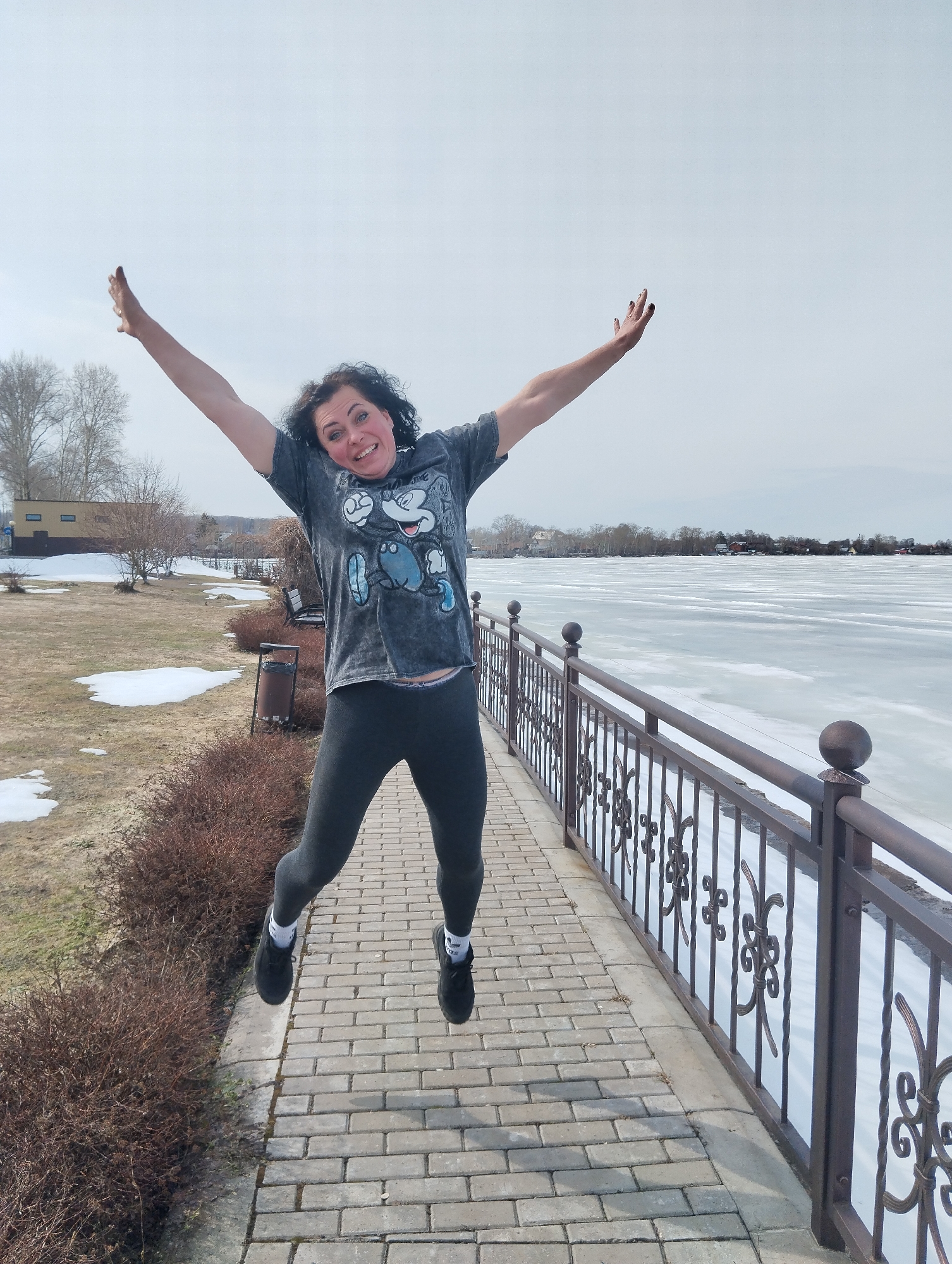 A woman is jumping mid-air with arms extended upward on a brick-paved path in Votkinsk, Russia. She wears a gray t-shirt with a graphic image, gray leggings, black shoes, and white socks. The path is bordered by a metal railing on the right, and vegetation on the left with patches of snow. Behind the railing is a frozen lake or body of water. In the background, there are buildings and trees, suggesting an urban landscape. The sky is overcast.