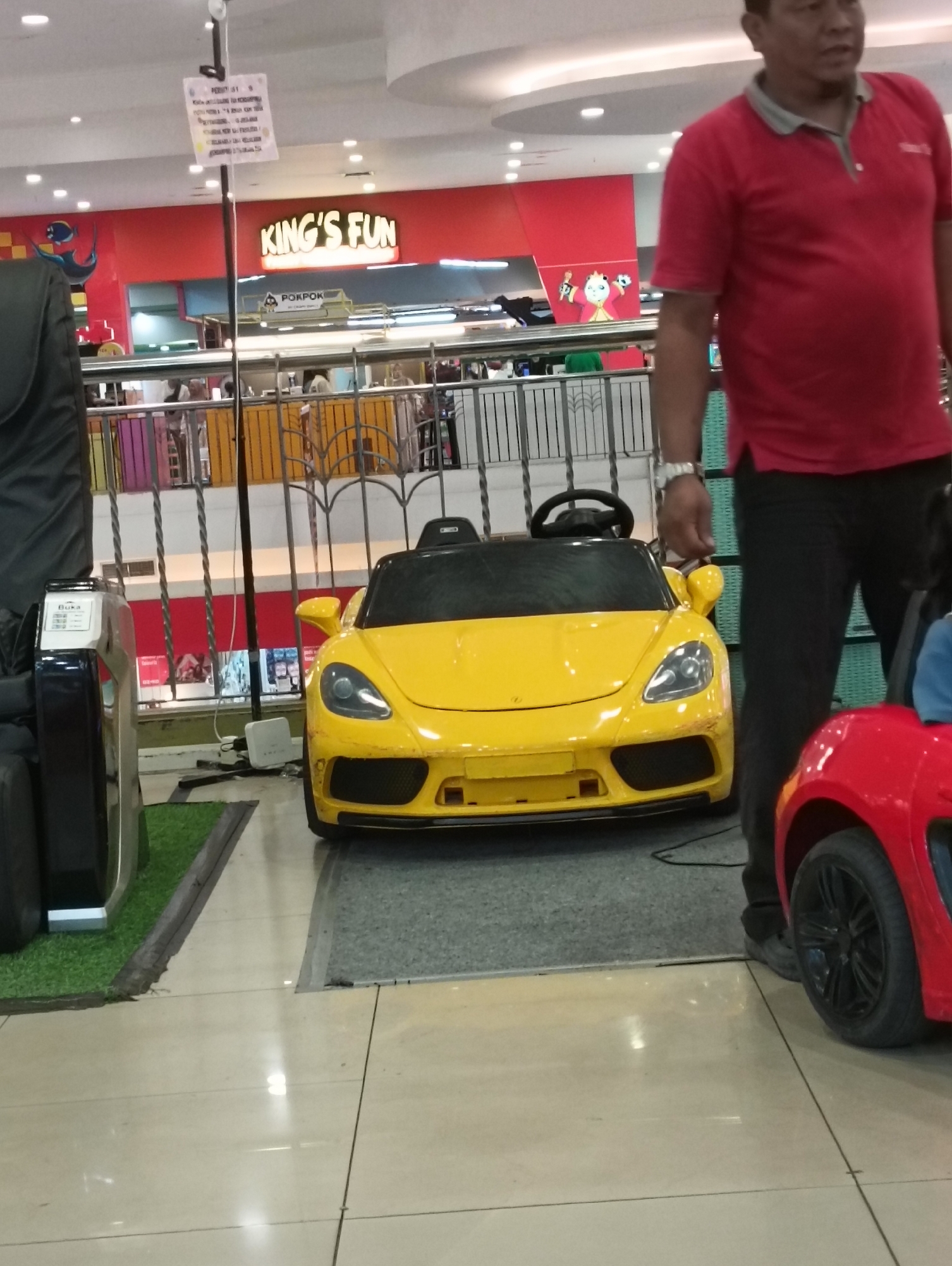 In a mall in Kediri City, Indonesia, a man in a red polo shirt and black pants stands near a yellow toy car. The toy car, resembling a sports car, is parked on a gray mat in front of a railing. Behind the railing, there's a store with a prominent "KING'S FUN" sign. A portion of a red toy car is visible to the right of the yellow car, and a black massage chair is on the left. The flooring is tiled and reflective. Above, there are ceiling lights and a hanging sign with text.