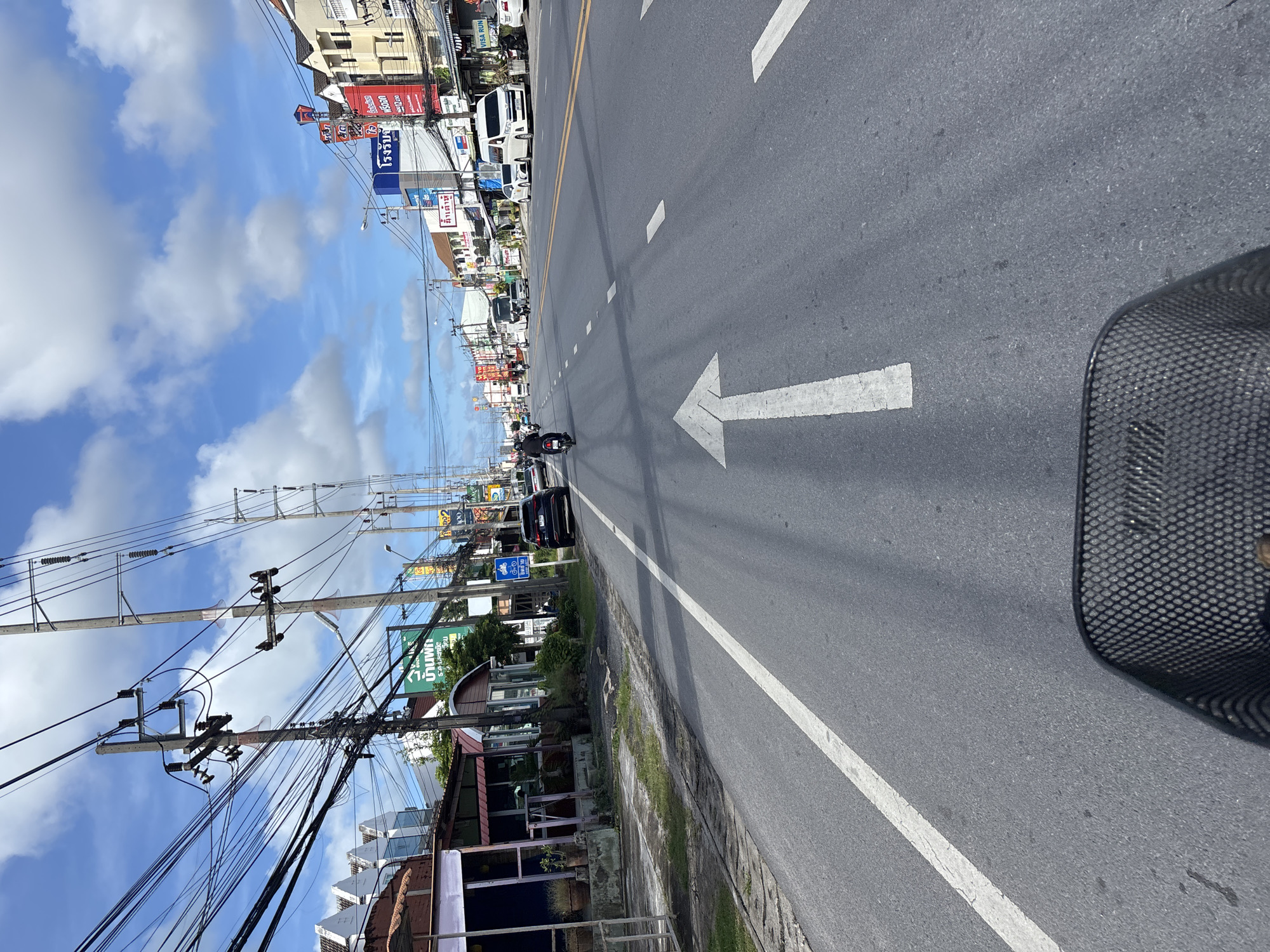 The image shows a street scene in Chalong, Thailand, on a partly cloudy day. The perspective is from a basket, likely on a bicycle or scooter, looking forward down a paved road with double yellow lines marking the center. A large white arrow indicates a forward direction. On the left side of the road, there are buildings with awnings and potted plants, alongside utility poles with a complex network of wires. On the right side of the road, more buildings are visible with various signs, including some in Thai. Traffic includes a dark SUV, a motorcycle with a rider, and several vans. The overall impression is of a busy, urban street in a tropical setting.