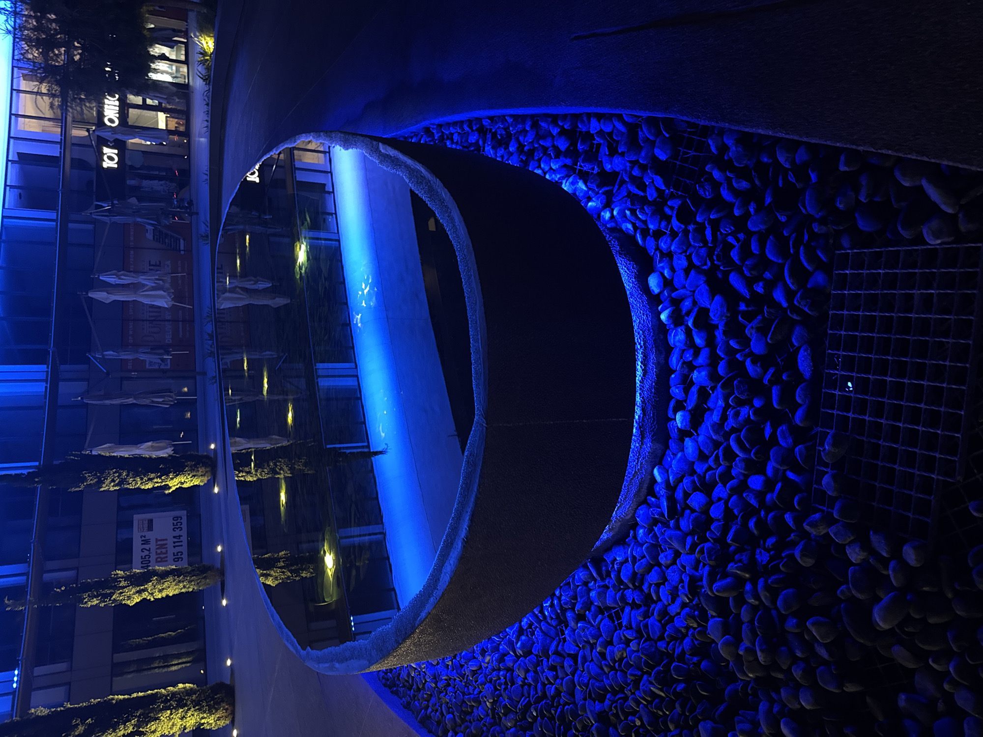 A reflective, dark grey, oval-shaped water feature is centrally located in the foreground of the image, illuminated by a vibrant blue light from within its base. The water surface reflects the surrounding environment, including the dark sky and building lights. The water feature is surrounded by a dense collection of dark grey and black river stones, also lit by the blue illumination. In the background, a modern building with large windows is visible, with blue lighting accentuating its facade. Trees with thin trunks and dark foliage stand tall on either side of the building. A white sign with black text reading "405,2 M² RENT 95 114 359" is attached to the building. Several closed white umbrellas are also visible near the building entrance. The scene is set at night, with the blue lighting creating a dramatic and somewhat abstract atmosphere. The location is identified as Germasogeia Municipality, Cyprus.