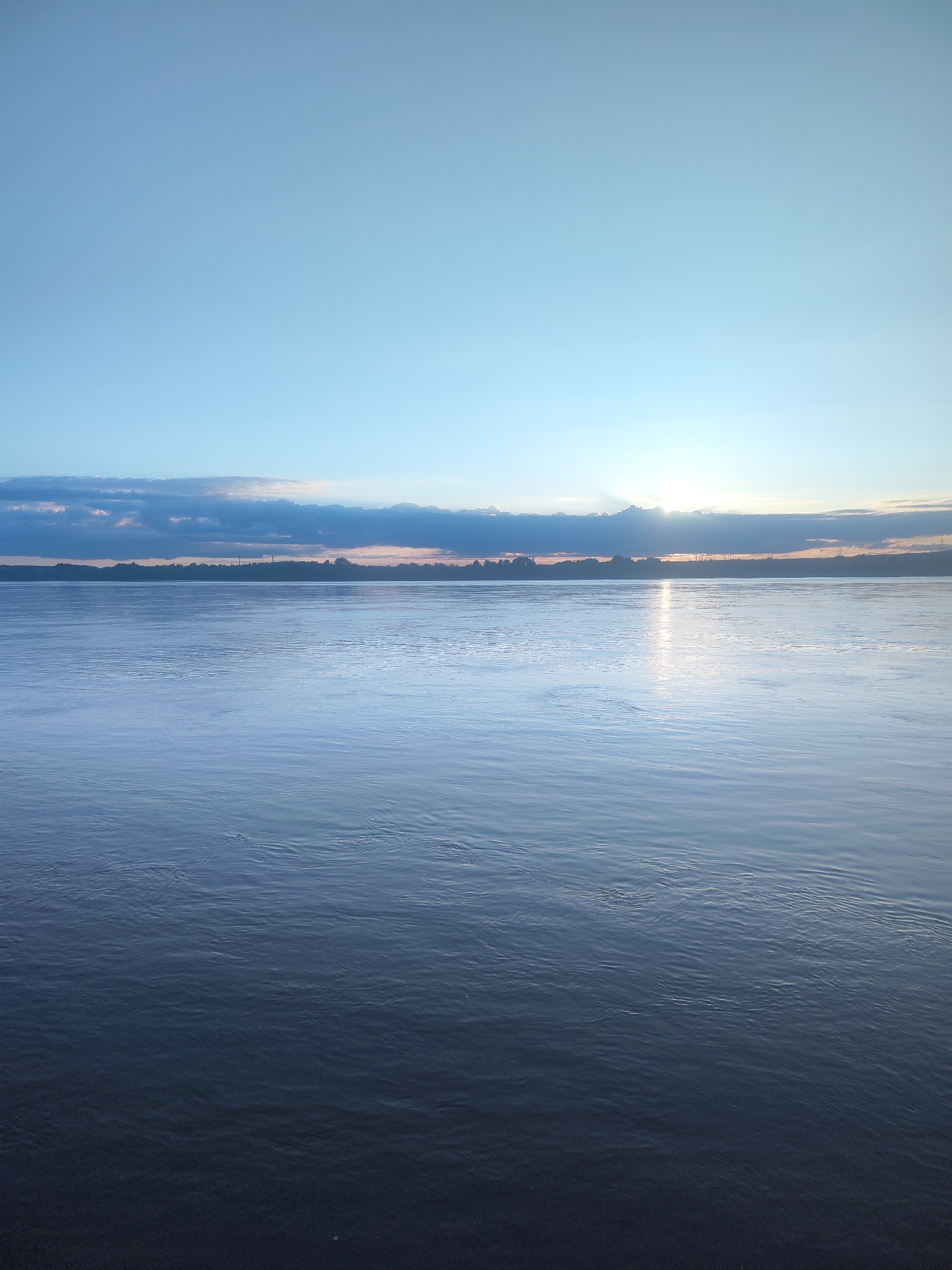 The image captures a serene waterscape at either sunrise or sunset, dominated by a wide expanse of water stretching towards a distant horizon. The sky above is a clear, light blue, transitioning to a slightly darker shade higher up. A prominent band of dark, horizontal clouds sits low on the horizon, partially obscuring the sun. The sun's rays pierce through a gap in these clouds on the right side, creating a bright reflection that shimmers across the water's surface towards the viewer.
On the distant horizon, a faint strip of land is visible, featuring a line of trees and what appear to be distant industrial structures or power line towers, silhouetted against the softer light. The body of water, likely a large river or lake, displays gentle ripples and reflections, with its surface appearing a cool blue and dark grey, deepening in color towards the foreground. There are no people, animals, or specific objects like boats clearly visible. The scene evokes a sense of peacefulness and vastness. No text is visible in the image.
