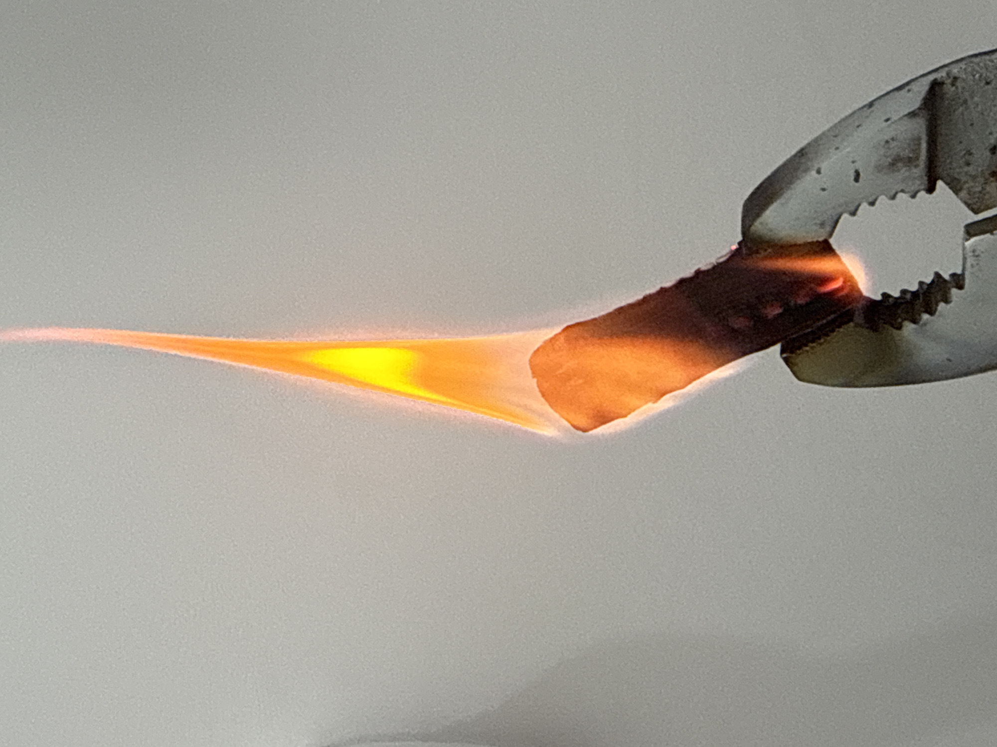 A lit roll of dark brown paper is held by a pair of tarnished metal pliers against a plain grey-white background. The paper is intensely lit with a yellow and orange flame that extends from the paper’s edge, narrowing to a point as it spreads leftward. The pliers grip the paper on the right, obscuring the edge. The pliers are grey, with visible wear and spots of rust. The background is featureless with a slightly textured surface. The overall lighting is bright, highlighting the fire and the textures of the objects. There is no indication of this being in San Bernardino Tlaxcalancingo, Mexico.