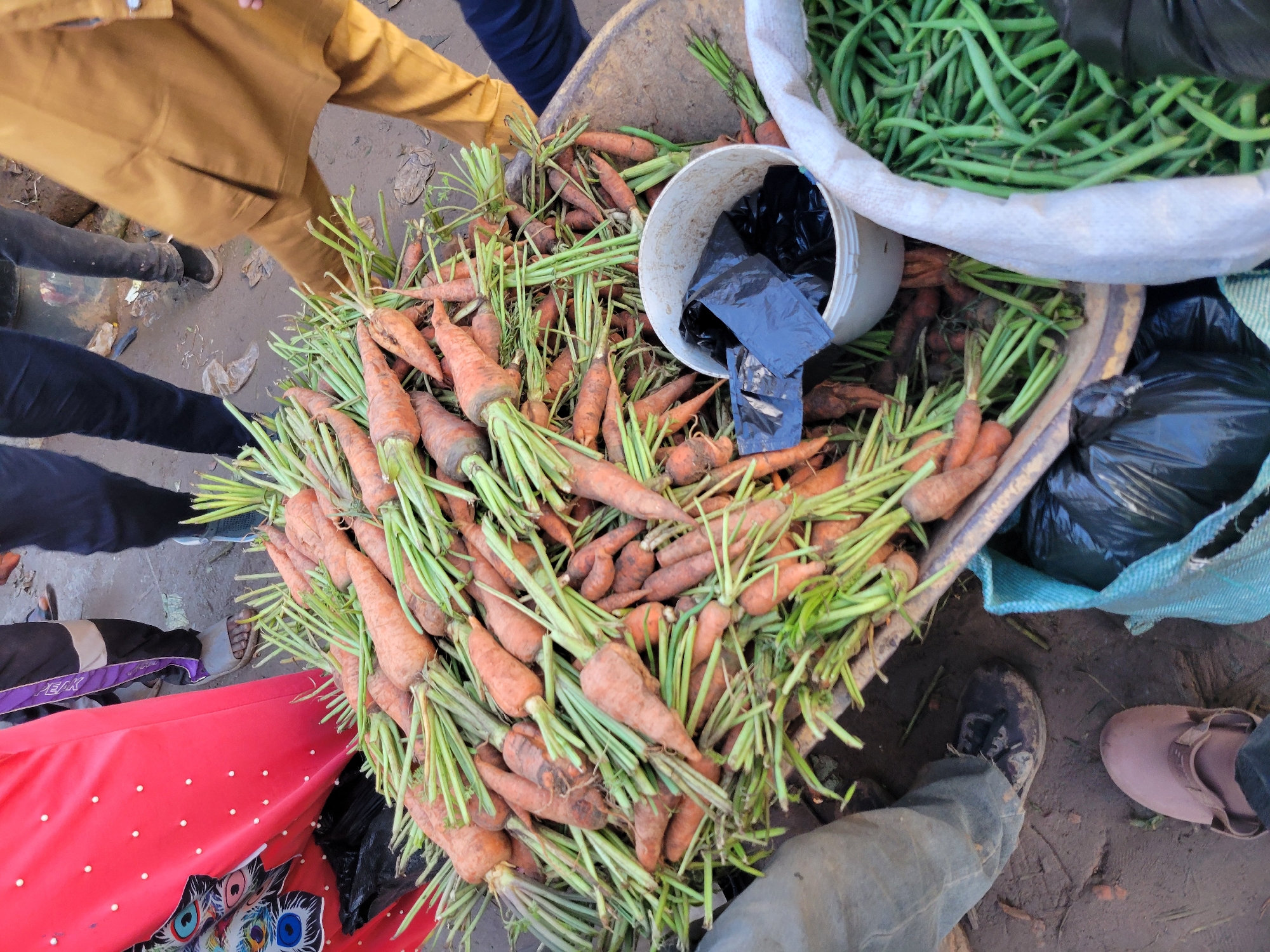 A street scene in Gura Top, Nigeria, depicts a market stall with a large pile of fresh carrots in a wheelbarrow. The carrots are orange with green tops, and appear to be of various sizes. Next to the wheelbarrow, a person wearing a brown tunic and pants is visible, as is another person wearing dark pants and shoes. In the foreground, a blue bag with black plastic bags inside is also present. The ground is muddy, suggesting recent rain or a poorly maintained market area. The overall impression is one of a bustling, informal market environment.