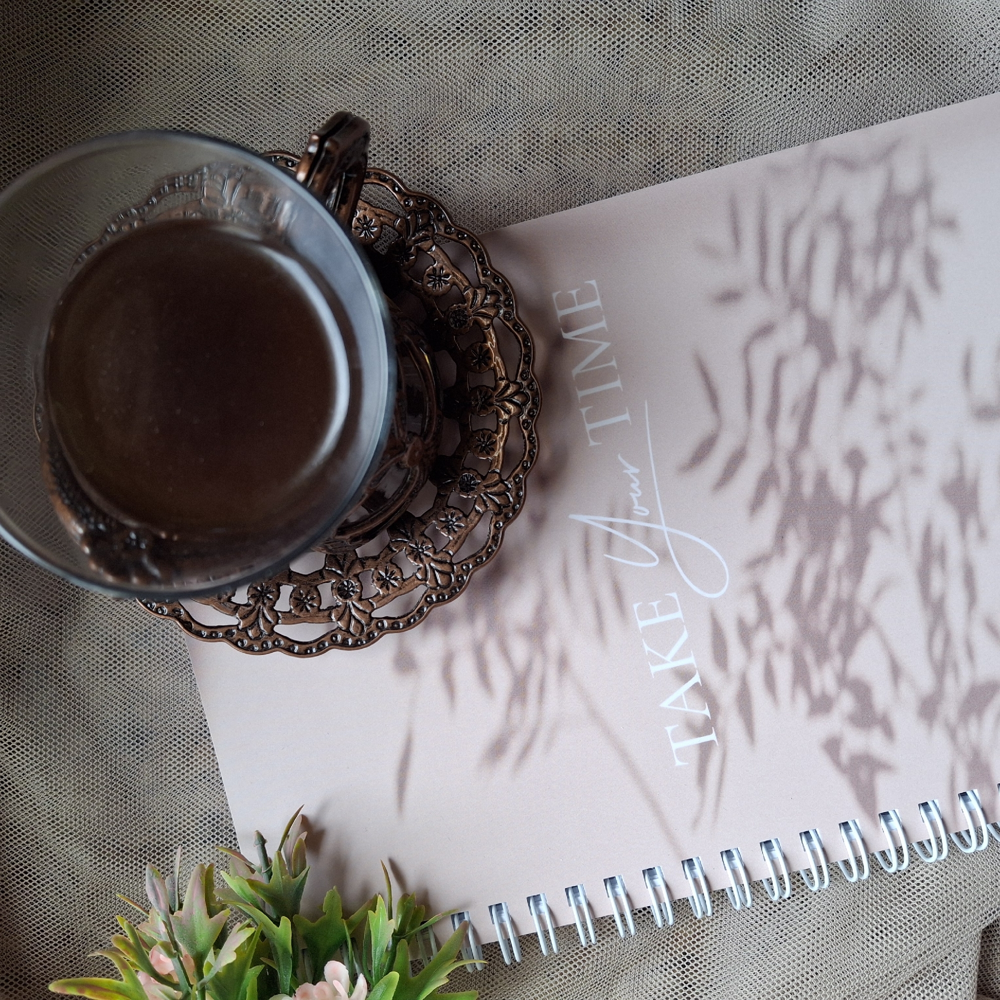 The image depicts a top-down view of a table setting. A glass cup filled with a dark liquid, possibly coffee or tea, rests on an ornate metal saucer. The saucer has a floral design with cut-out spaces. Next to the cup is a spiral-bound notebook with a pale pink cover. The words "TAKE Your TIME" are printed on the cover in white. Leafy shadows are cast across the notebook. In the lower left corner, there is a small arrangement of artificial flowers with green leaves and light pink blossoms. The entire scene is set on a textured beige fabric surface. The location is Unknown, Unknown.