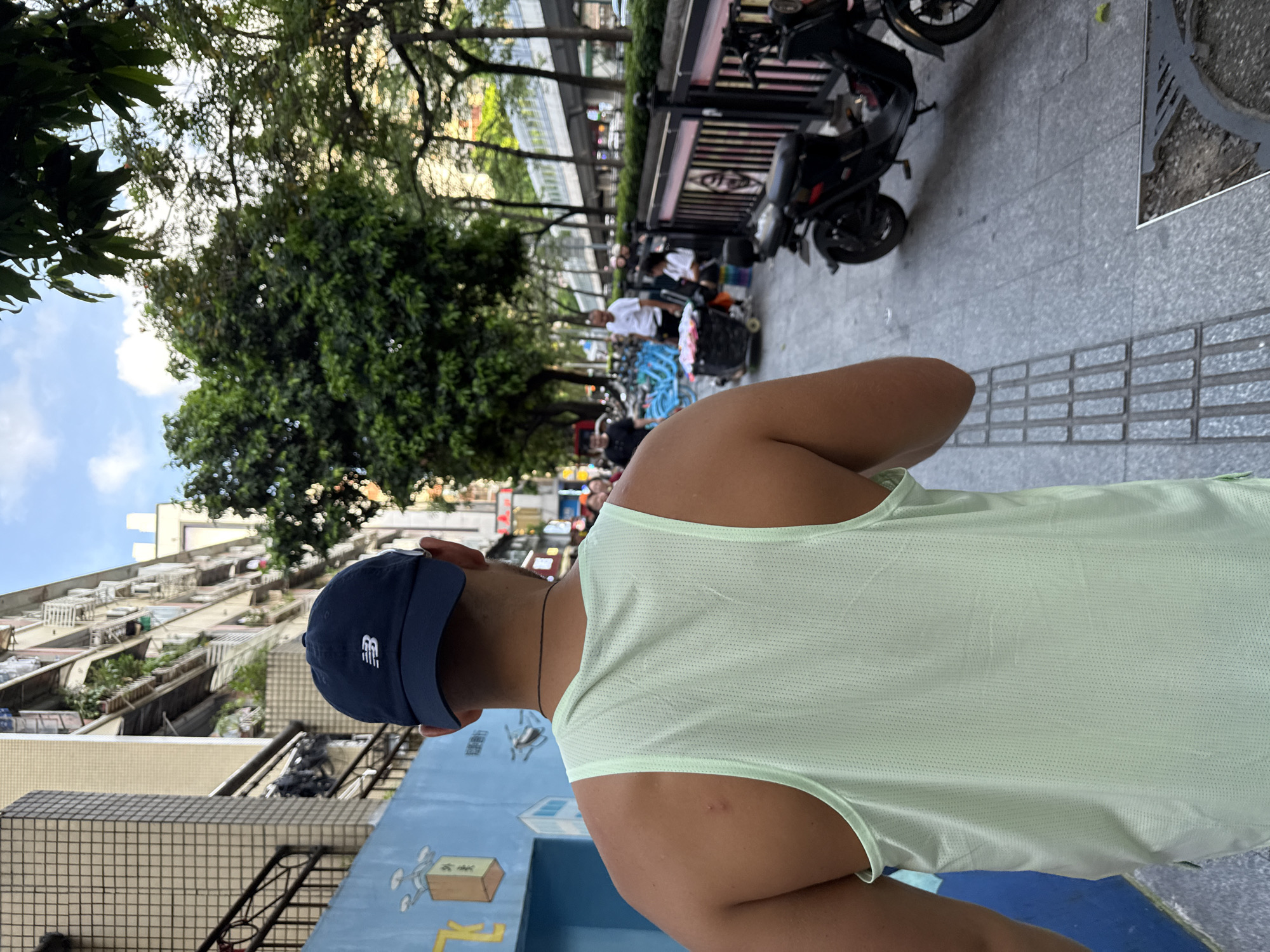 A man wearing a light green tank top and a navy blue baseball cap walks down a sidewalk in Haizhu District, China. The man is viewed from behind, and his back and shoulders are visible. The sidewalk is paved with grey square tiles and has a tactile paving strip running along its edge. To the left of the sidewalk is a blue wall with drawings on it. Behind the wall, buildings with multiple floors and balconies are visible. To the right of the sidewalk is a metal fence and trees. Several bicycles are parked behind the fence, and a black electric scooter is parked on the sidewalk. In the background, there are more trees and a raised pedestrian walkway with traffic on it. Several people are visible walking and standing in the background. The sky is blue with some clouds.