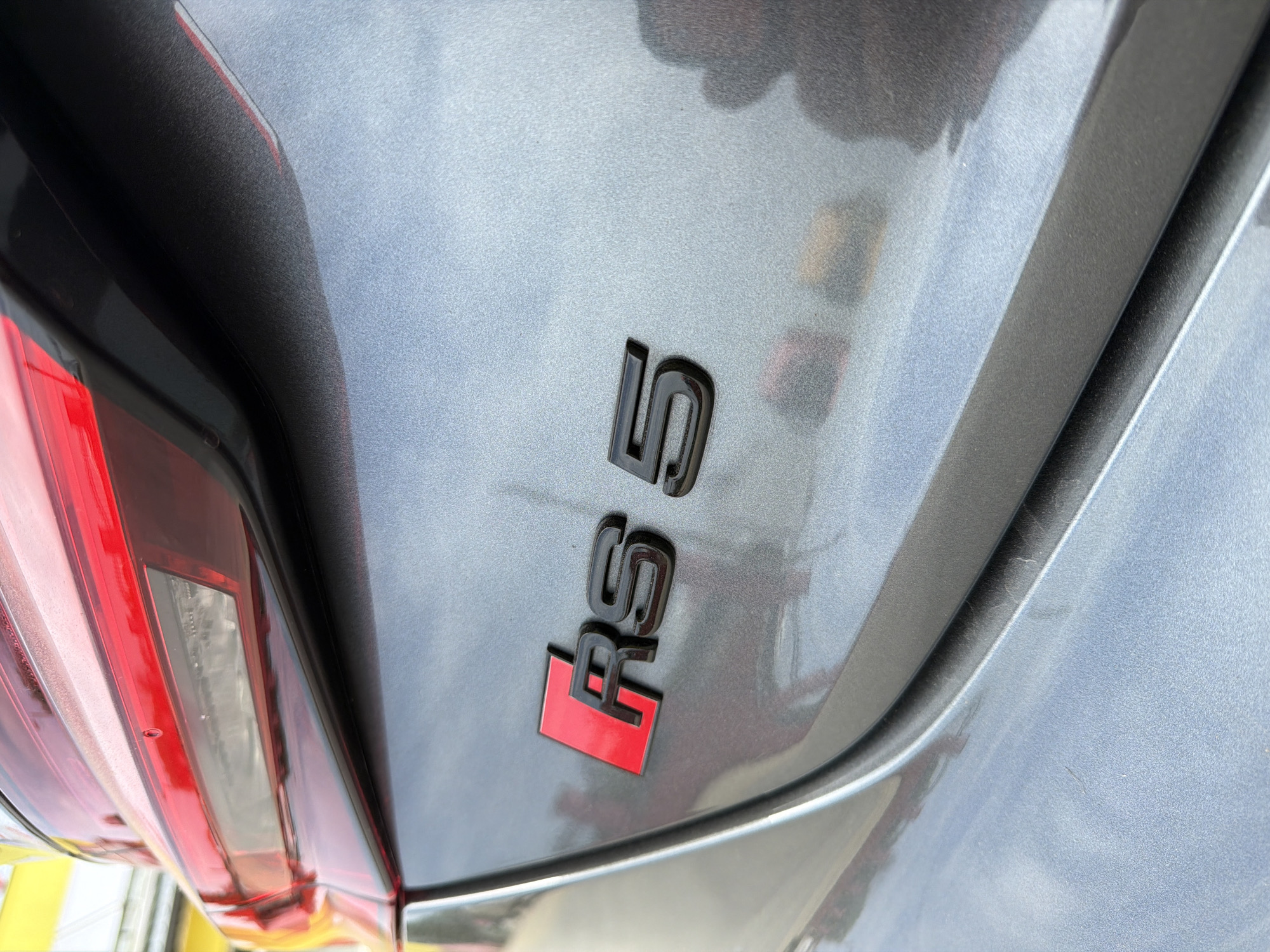 The image shows the rear of a gray Audi RS 5. The "RS 5" emblem is visible on the right side of the frame. The left side of the image shows the car's tail light and bumper. No other objects or people are visible. The location cannot be determined from this image.