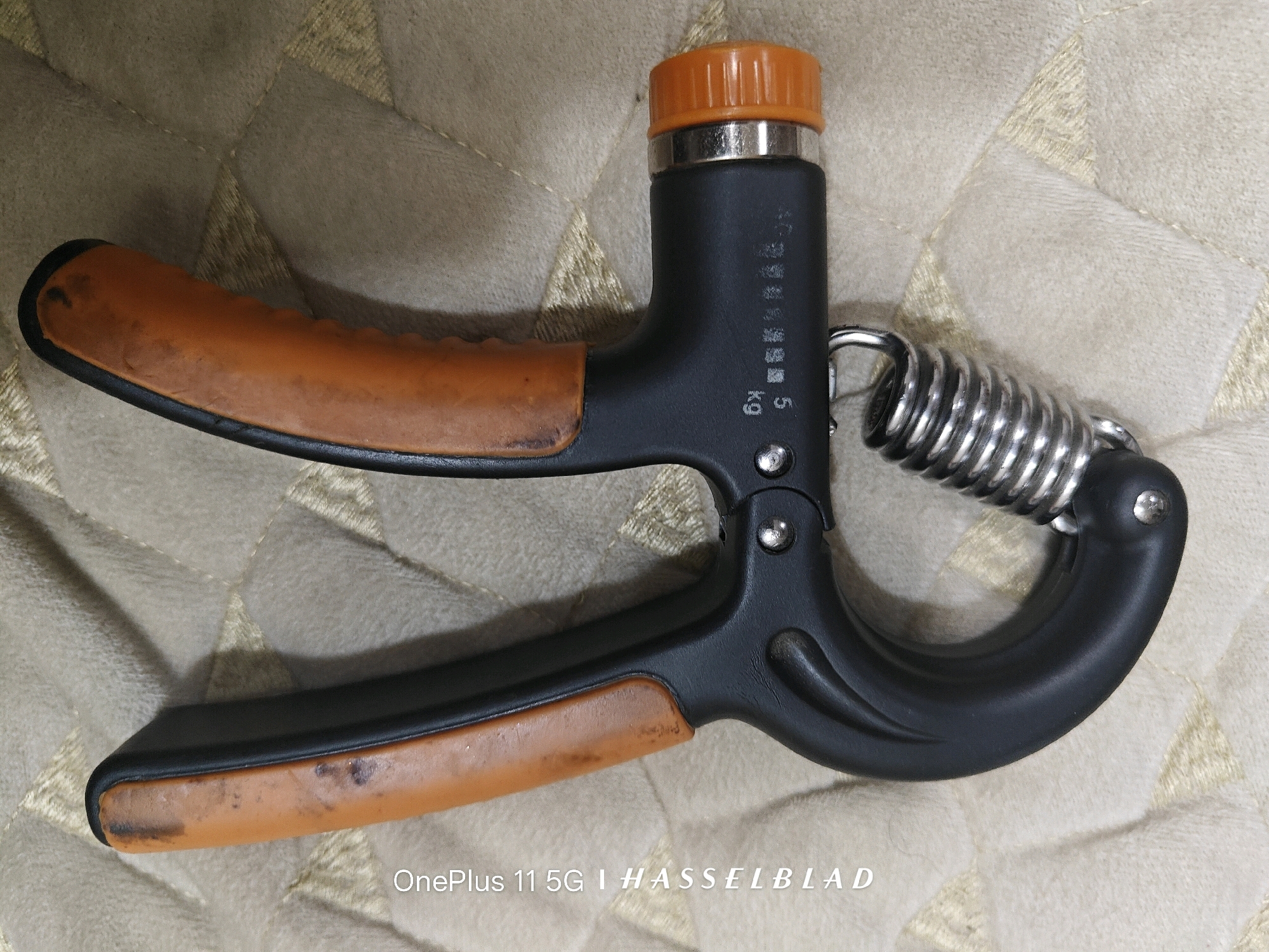 A black and orange hand gripper is placed on a textured, beige surface, possibly a carpet or fabric. The gripper has two handles, one orange and the other black with an orange grip, connected by a silver spring. There is some writing on the black handle that appears to be in Chinese characters and the number "5" followed by "kg". The bottom right corner of the image has a watermark that reads "OnePlus 11 5G | HASSELBLAD". The lighting suggests an indoor setting. There are no people present in the image, nor are there any indications of a specific location within Nagpur, India.