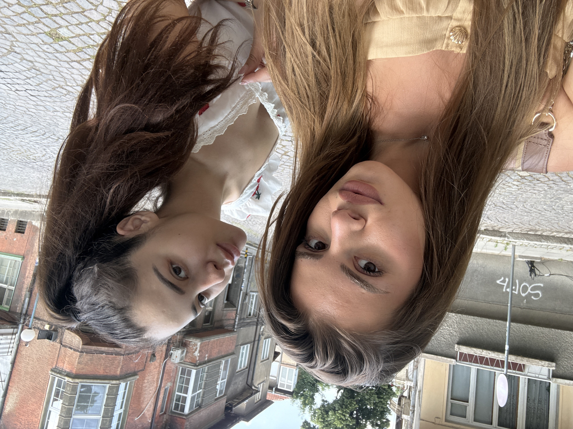 The image, when correctly oriented (rotated 90 degrees counter-clockwise), captures two young women positioned closely together, looking directly at the camera, likely in a selfie-style shot. The scene takes place outdoors during the daytime, under bright, possibly overcast, conditions, in what appears to be an urban environment consistent with Plovdiv, Bulgaria.
The woman on the left has long, dark brown hair and fair skin. She is dressed in a white top featuring red embroidered accents and a delicate lace trim along the neckline. Her expression is neutral.
The woman on the right has long, lighter brown or auburn hair and fair skin. She wears a light beige or tan top characterized by a ruffled neckline and a distinctive golden, shell-shaped button. A delicate silver chain with a small, round pendant adorns her neck. Her expression is also neutral, with full lips.
The background showcases old European urban architecture. Visible structures include multi-story buildings constructed from brick and plaster, featuring numerous windows, some of which are bay windows. The ground beneath them is paved with cobblestones. Green foliage from trees is visible in the lower right area of the frame. On a grey wall in the upper right background, white text or graffiti can be seen, appearing to read "4.JOS" or "4.IOS". The overall mood of the image is casual and observational.