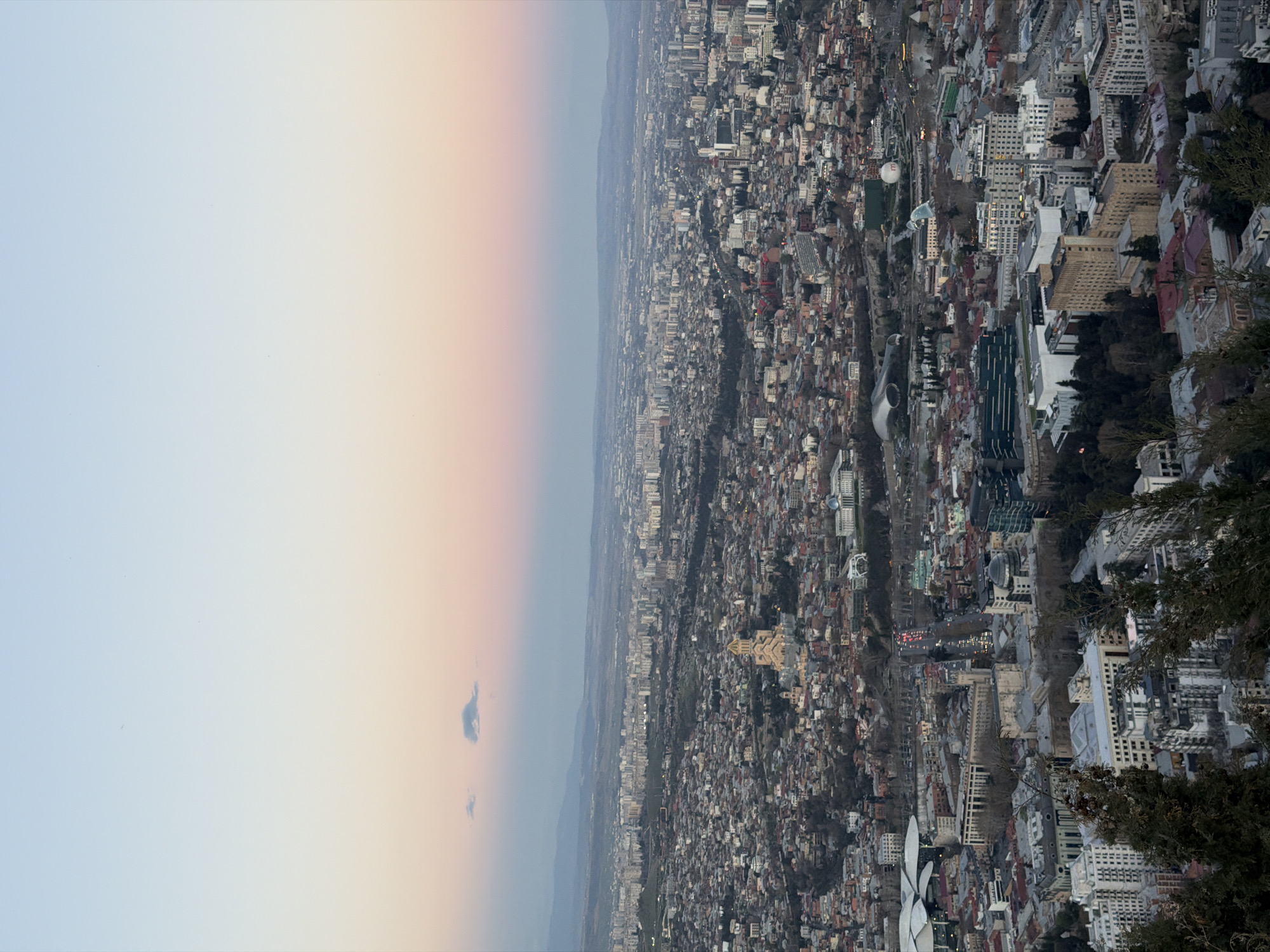 The image depicts an aerial view of Tbilisi, Georgia, likely taken at sunset or sunrise given the gradient sky. The city is densely populated with a variety of buildings, ranging from residential houses to high-rise apartments. A river or valley appears to cut through the cityscape. Some greenery is visible, indicating parks or wooded areas. The shot seems to have been taken from a high vantage point, possibly a mountain or tall structure, with some trees or foliage in the lower right corner of the frame.