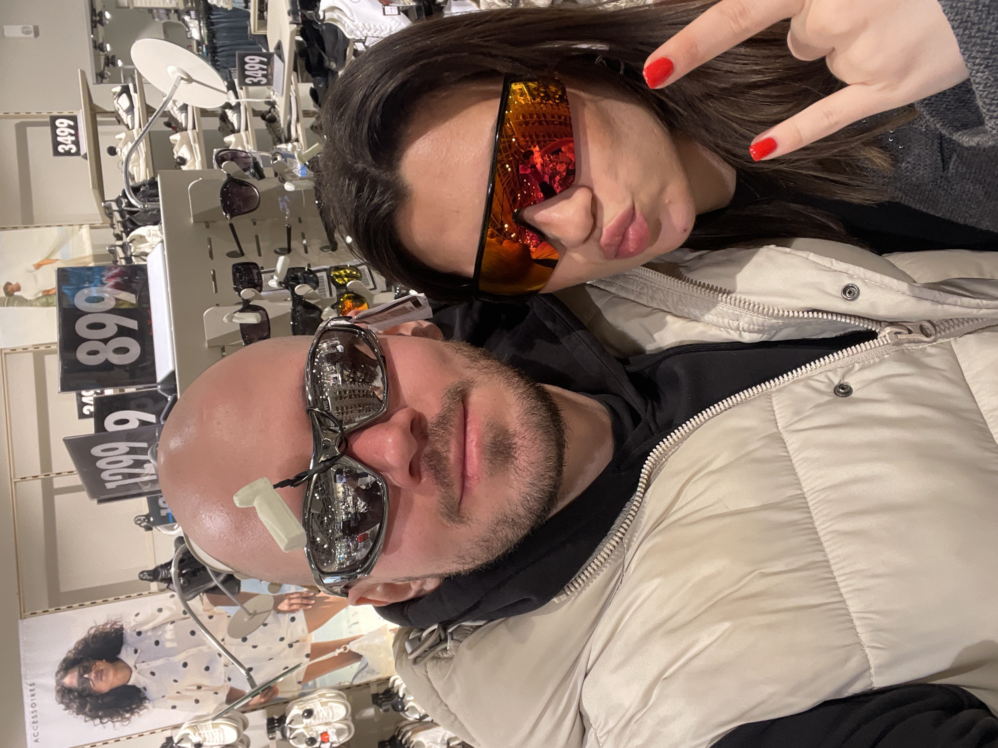 The image shows a selfie of two people indoors. A bald man wearing reflective silver sunglasses and a beige puffer vest over a black hoodie is in the foreground. He has a white bandage on his forehead near the temple. Behind him is a woman with long brown hair, wearing orange reflective sunglasses and a grey sweater, making a rock-and-roll hand gesture. The background shows a display of sunglasses and price tags. A poster with a woman wearing glasses and a polka-dot dress is visible. The environment suggests a retail store, possibly a clothing or accessories shop, with organized displays. The location is specified as Krasnoyarsk, Russia, although this is not visually verifiable from the image alone.