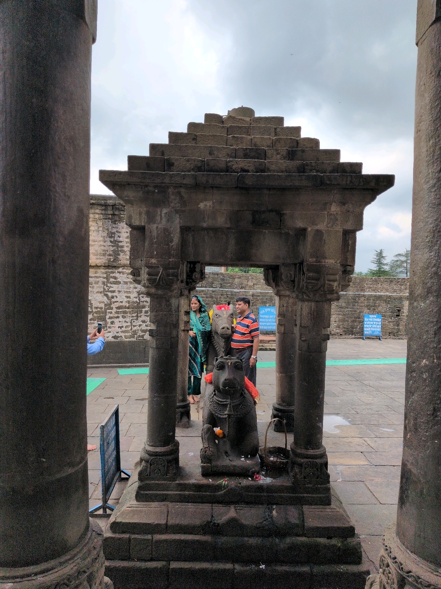 In the city of Baijnath, India, within an outdoor courtyard paved with stone tiles, a stone structure with a tiered roof sits prominently in the center. This structure is supported by four large stone pillars and houses a dark stone statue of an animal, possibly a bull, adorned with a red cloth around its neck. Two people stand near this statue; a woman in a teal and dark blue patterned sari and a man in an orange and striped t-shirt. To the left of the central structure, a person in a blue shirt is captured taking a photograph of the scene with their mobile phone. In the background, a stone wall encircles the courtyard, and on the wall, two blue signs are affixed. The sky above is overcast with clouds.