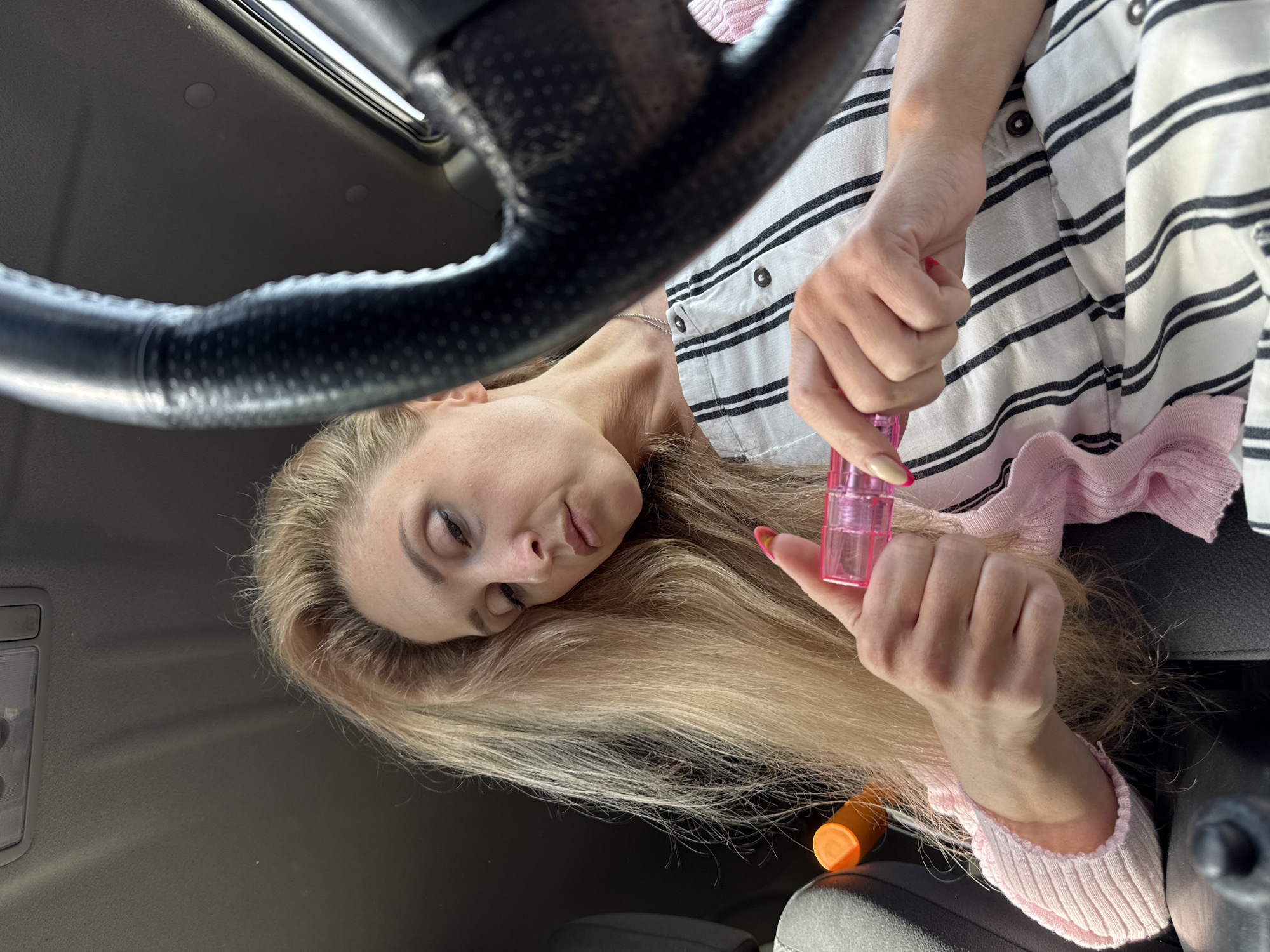 A blonde-haired woman is in the driver's seat of a car, holding a pink spray bottle. She is wearing a striped shirt with a pink sweater underneath. A steering wheel is visible on the left side of the image. The scene appears to be inside a vehicle, and the exact location is not discernible from the image. There is no indication or evidence in the image to confirm the location as Chaikovsky, Russia.