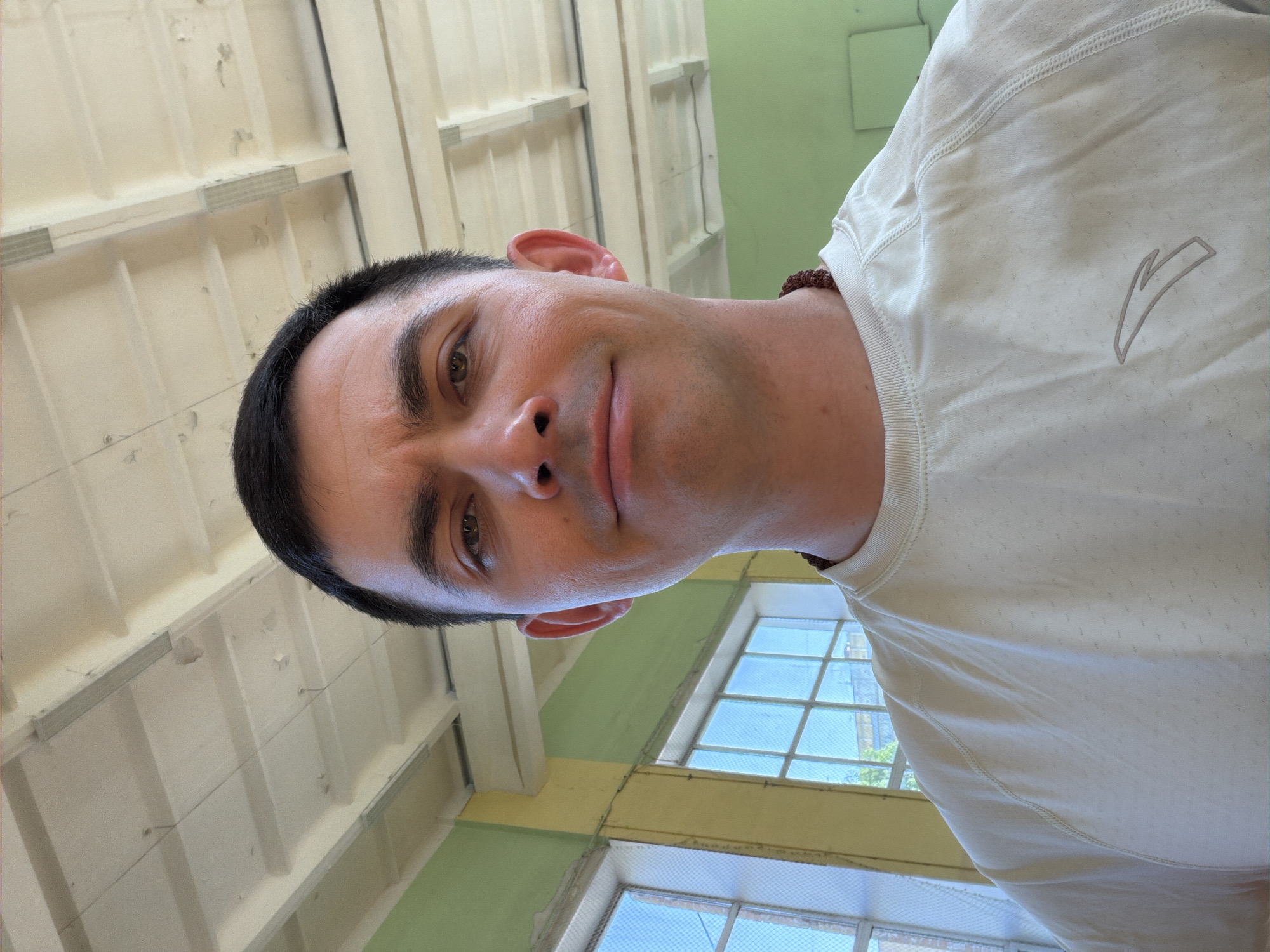 A medium, slightly rotated close-up of a fair-skinned man with short, dark hair and brown eyes. He is facing the camera with a slight smile. He is wearing a white athletic t-shirt with a logo on the upper right chest. Behind him, there is a light green wall and windows with light blue panes. The ceiling consists of white panels with darker beams. This could be inside a building in the city of Izhevsk, Russia, but there is no way to confirm location.