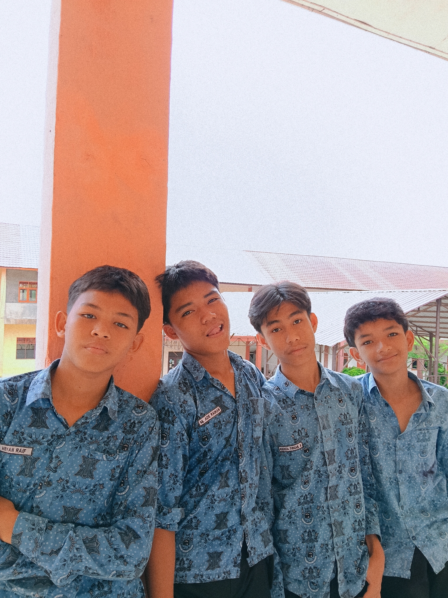 Four teenage males in blue patterned shirts pose for a photo against an orange and white column in Lhokseumawe, Indonesia. The shirts have name tags with the names "ABYAN RAIF", "M. ADE RAHMAN", and "RANGKUL TURGAK A." on them. A building is visible in the background.