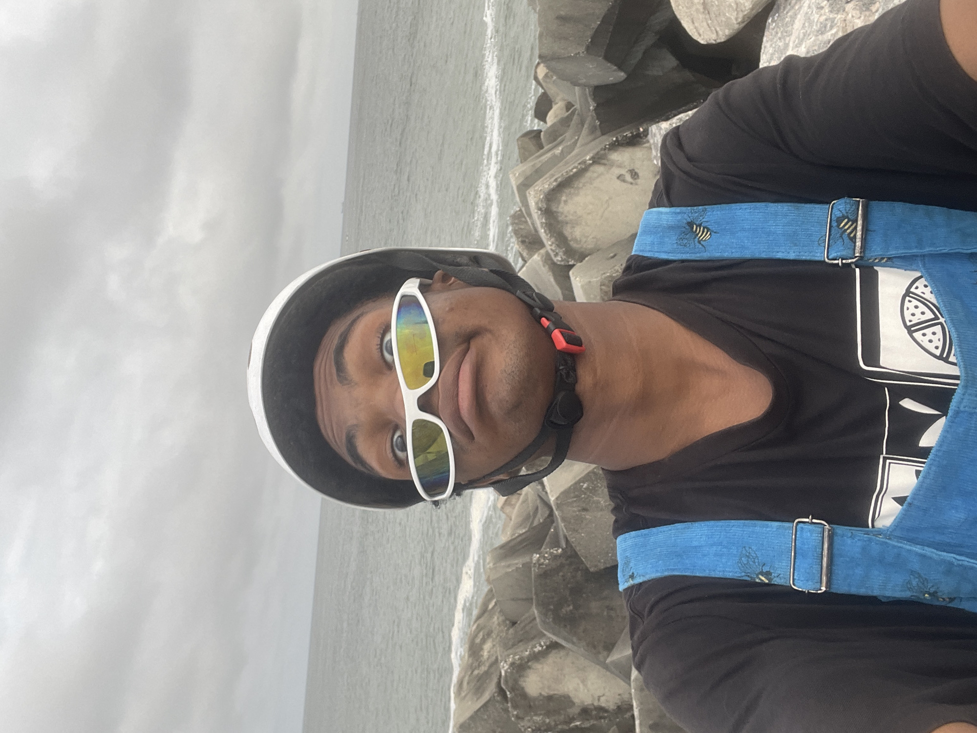 In Inupa Kekere, Nigeria, a young person wearing a white helmet, white wraparound sunglasses, and blue overalls over a black t-shirt takes a selfie by the ocean. The person is looking at the camera with a humorous expression, with one eye looking directly at the lens and the other looking slightly upward. The background shows a rough, rocky coastline with large concrete blocks, and the ocean extends to the horizon under a cloudy sky. The person's posture and facial expression suggest a playful or humorous mood.