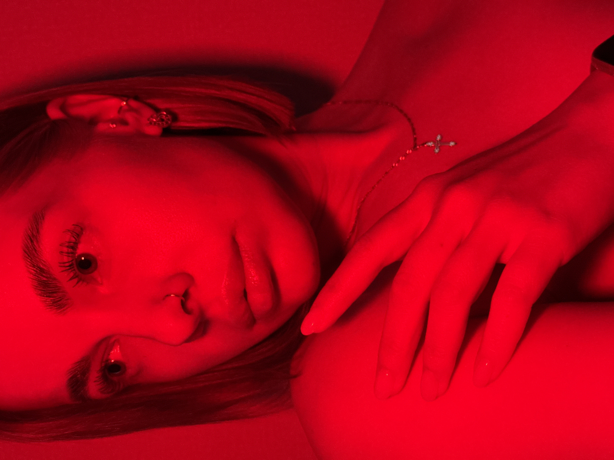 This close-up, angled shot features a young woman reclined, illuminated by intense red lighting. Her face, positioned towards the viewer, is the central focus, showcasing her defined eyebrows and long, dark eyelashes. She has dark eyes and her gaze appears direct and contemplative. A small nose ring (septum piercing) is visible, along with small earrings in her left ear—one hoop and one stud. Her lips are slightly parted.
Her light-colored hair is visible around her head, particularly on the left side of the frame. A delicate necklace with a small, sparkly cross pendant rests on her chest, and a dark band, possibly a watch or bracelet, is on her right wrist. Her left hand, with manicured fingers, rests gently on her upper body.
The entire scene is bathed in a saturated red light, casting deep shadows and strong highlights that emphasize the contours of her face and body. This monochromatic red illumination creates a dramatic, intense, and somewhat mysterious atmosphere. There are no discernible elements of clothing, and the background is an abstract, uniformly red environment, suggesting an indoor or studio setting. No text is visible in the image. The activity is a posed portrait.