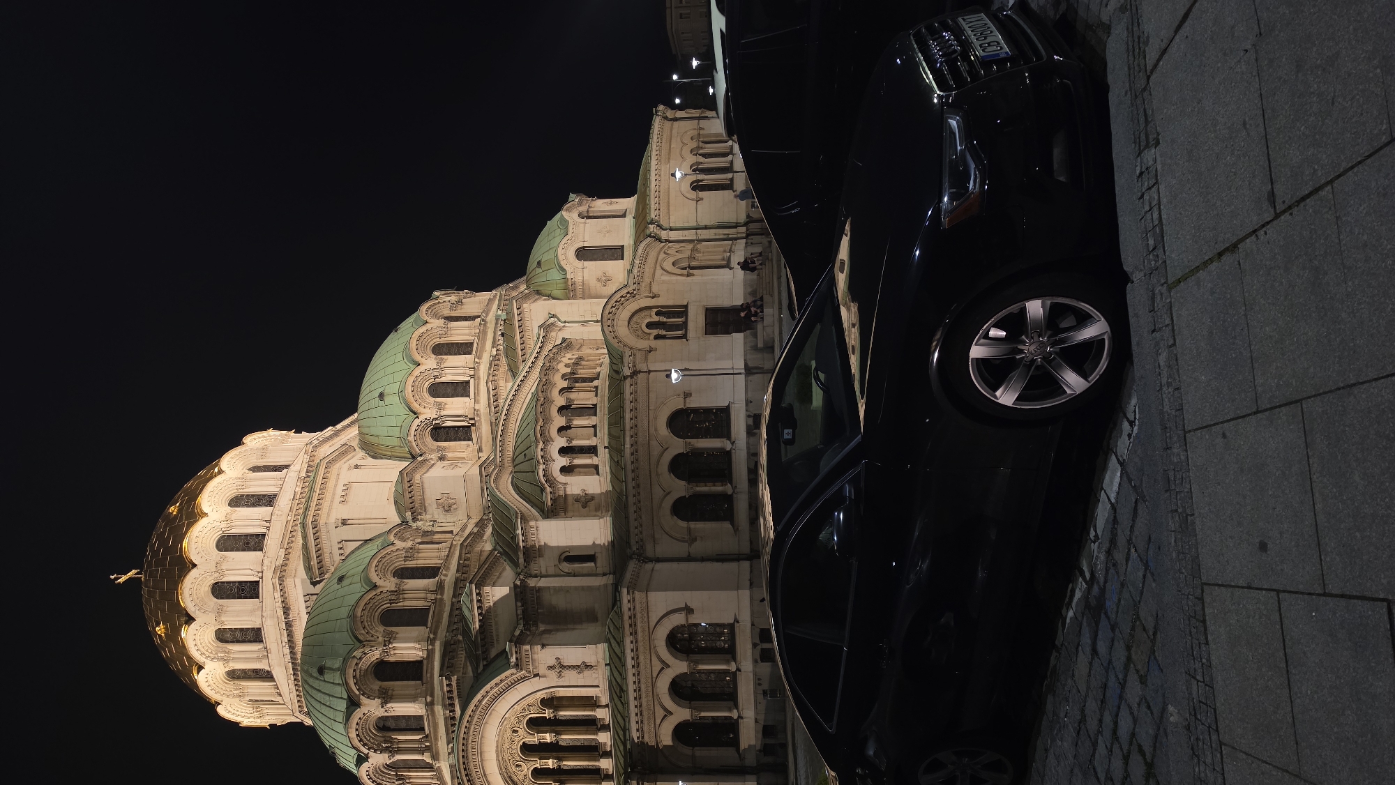 The image presents a striking nighttime view of the Alexander Nevsky Cathedral in Sofia, Bulgaria, partially obscured by two parked cars. The majestic cathedral, situated on the left and center, is brightly illuminated against a dark, clear night sky. Its distinctive features include several large, green-patinated domes and one prominent golden dome, which shines brilliantly, topped with a cross. The building's ornate facade showcases intricate light-colored stonework, multiple tiers, and numerous arched windows, all highlighted by artificial lighting that emphasizes its architectural grandeur.
In the foreground and to the right, two dark-colored vehicles are parked on a stone-paved surface. The closest car appears to be a black Audi, with its front wheel, side, and part of its front visible. A license plate on this car clearly displays "A 0086 XT". Another dark vehicle is parked behind it. The ground, visible beneath and around the cars, is made of grey, rectangular stone paving blocks. No discernible activity or people are visible, contributing to a serene and monumental atmosphere. The scene captures the iconic cathedral's beauty under night illumination.